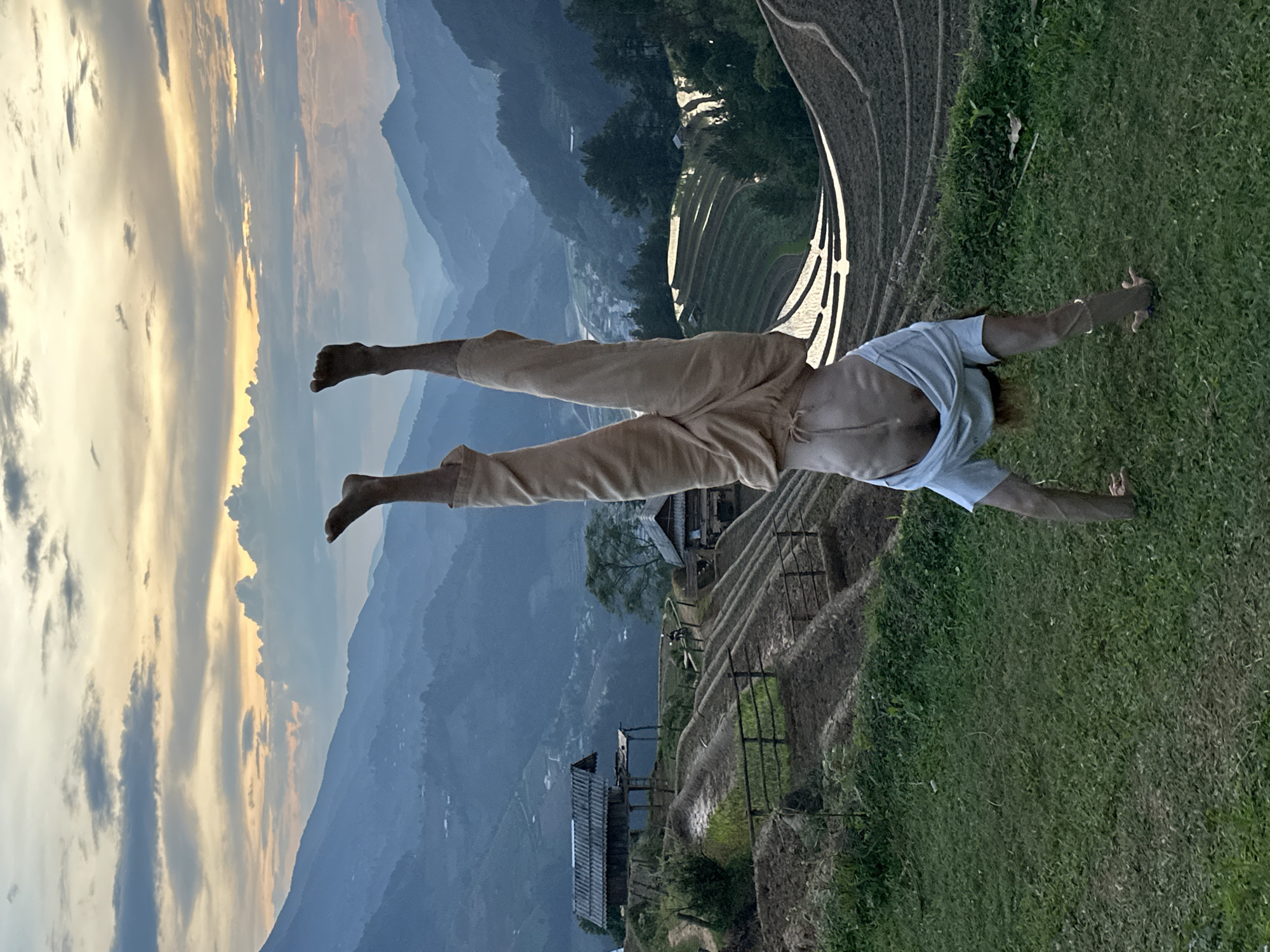 A person is captured in an acrobatic or yoga pose, appearing to float or fly, against a dramatic mountainous backdrop. The individual, an adult, is lying prone with arms extended forward and slightly outward, and legs extended backward, both lifted off the ground. They are dressed in light brown pants and a light blue short-sleeved t-shirt.
The scene is set in a vast mountainous landscape, likely Mù Cang Chải, Vietnam, characterized by intricately carved rice terraces that wind along the hillsides, particularly prominent on the right. Small, traditional buildings and scattered trees are visible among the terraces and in the valleys below. Layers of mountains, hazy blue in the distance, form the far background.
The sky, occupying the upper left quadrant, displays warm, golden, and orange tones merging with softer blues and grays, indicating either sunrise or sunset. Clouds are scattered across the sky, catching the warm light. The immediate foreground consists of a lush green grassy area, from which the person is performing their pose, overlooking the expansive vista. The image conveys a sense of freedom, athleticism, and connection to the natural, cultivated environment. No visible text is present.