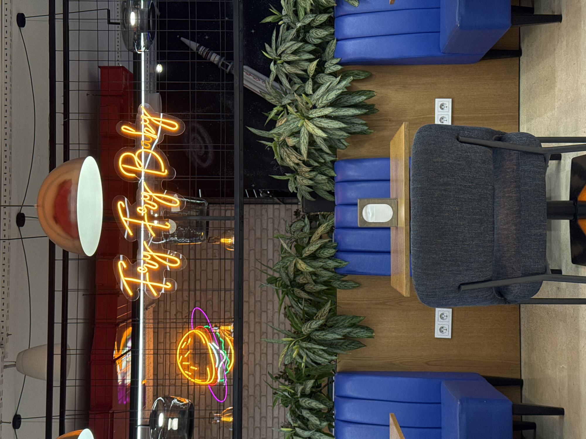 The image depicts an interior space in Yerevan, Armenia. The view is from above, showing a seating arrangement with blue cushioned benches on either side of a wooden table. A gray upholstered chair is positioned on the right side of the table. Decorative plants are placed along the center, between the benches and the table.
On the left side of the frame, there is a black metal grid structure featuring various decorative lighting elements, including a large circular lamp and a neon sign. The backdrop behind the grid includes a mural or wall art featuring space-themed imagery, such as a rocket and stars. The environment suggests a modern, stylized interior, possibly a cafe or restaurant. There are electrical outlets visible on the wooden paneling behind the benches.
