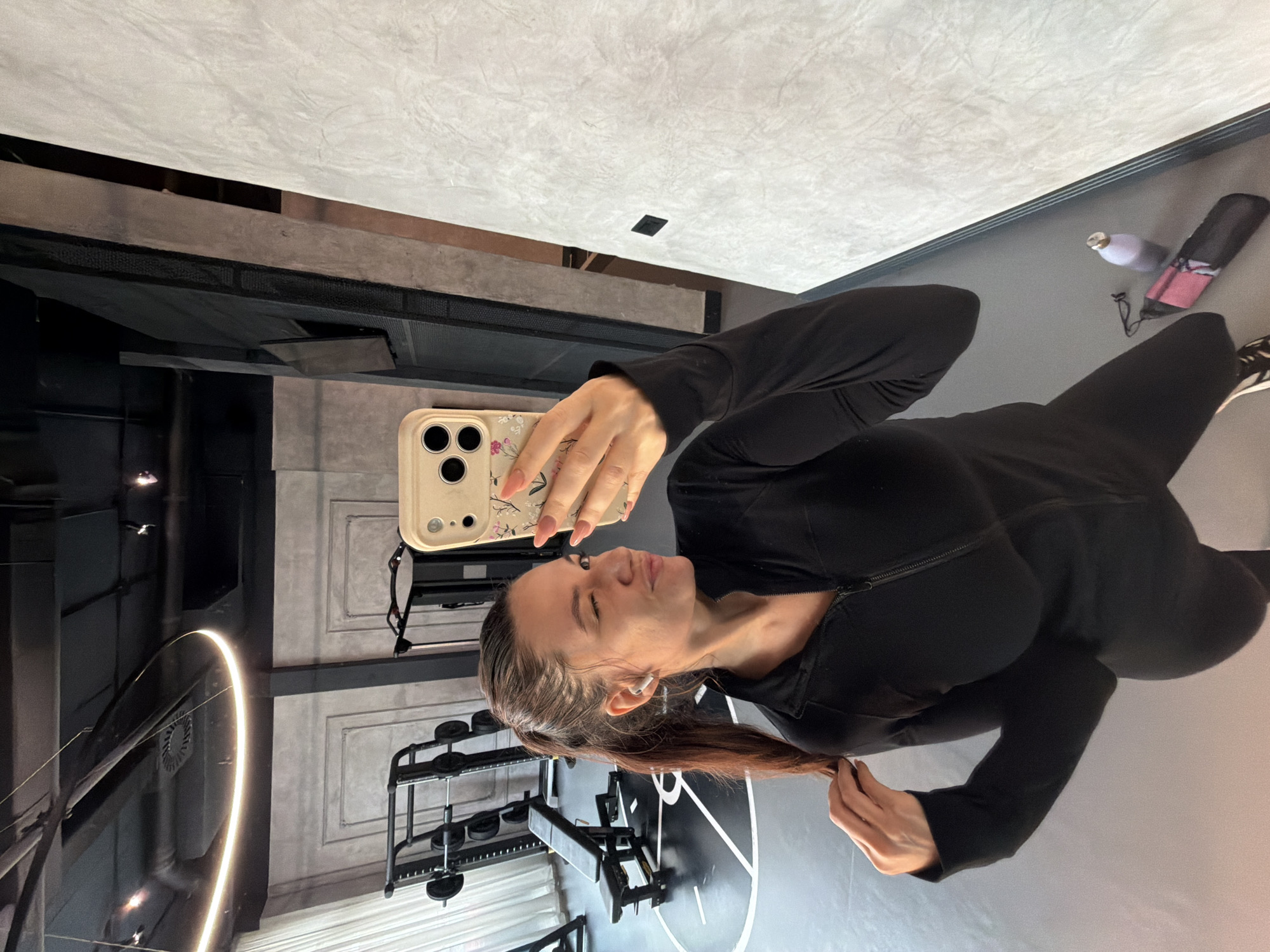 A woman in black athletic wear takes a selfie with a beige smartphone. The setting appears to be a gym in Belgrade, Serbia, indicated by Cyrillic script on a wall design (not visible in this crop). Gym equipment, including weight racks and benches, are visible in the background, along with yoga mat and water bottle. The lighting suggests a modern facility with ample natural and artificial light.