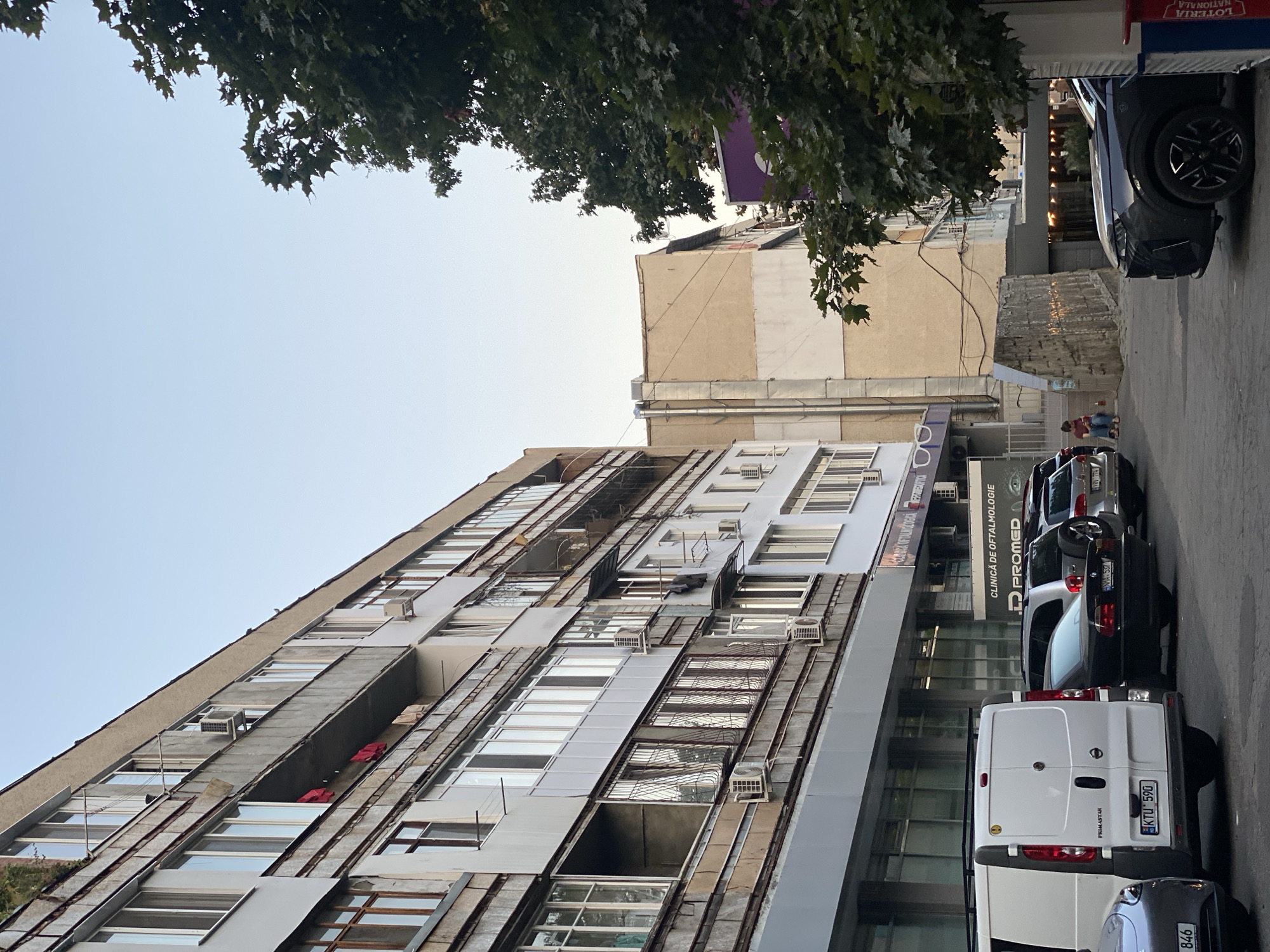 The image depicts a street scene in Chișinău, Moldova. In the foreground, a white van with the license plate KTU 590 is parked, partially obscuring a silver car. To the right of the van, a black sedan with the license plate NMA 537 is parked, followed by a silver SUV with a spare tire mounted on the back, and another silver car with the license plate YBY 774. A dark grey SUV is parked on the far right.
The buildings lining the street are multi-story structures. The building on the left is a weathered, concrete apartment building with multiple windows and balconies. Some balconies have laundry hanging out. Air conditioning units are visible on the exterior of the building. A sign above a ground-level establishment reads "CLINICĂ DE OFTALMOLOGIE" and "D PROMED."
Adjacent to this building, to the right, is a lighter-colored building with a more modern facade. Further to the right, a large tree with dark green leaves partially obstructs the view of the sky and the buildings behind it. A purple sign is partially visible behind the tree.
In the background, a person is seated on a low wall on the right side of the image. The sky above is a clear, pale blue. The overall impression is an urban environment with a mix of older and newer architecture.