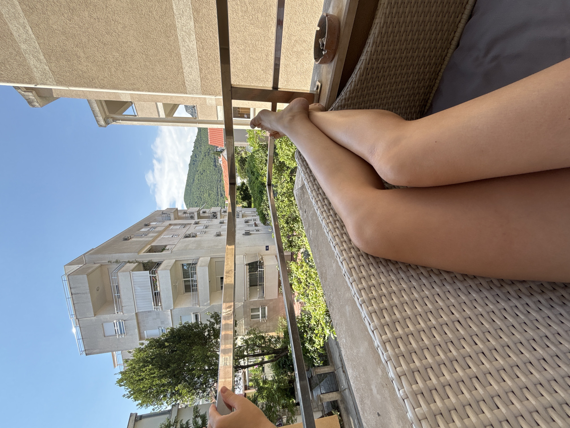 The image shows a person's legs crossed while sitting on a wicker chair on a balcony, with a view of buildings and a tree. The background depicts multi-story buildings with balconies, a green tree, and lush vegetation. In the distance, hills covered in trees are visible under a clear blue sky with some clouds. An ashtray with cigarette butts rests on the balcony railing. The specific location in Budva, Montenegro, cannot be determined from the visual information provided.