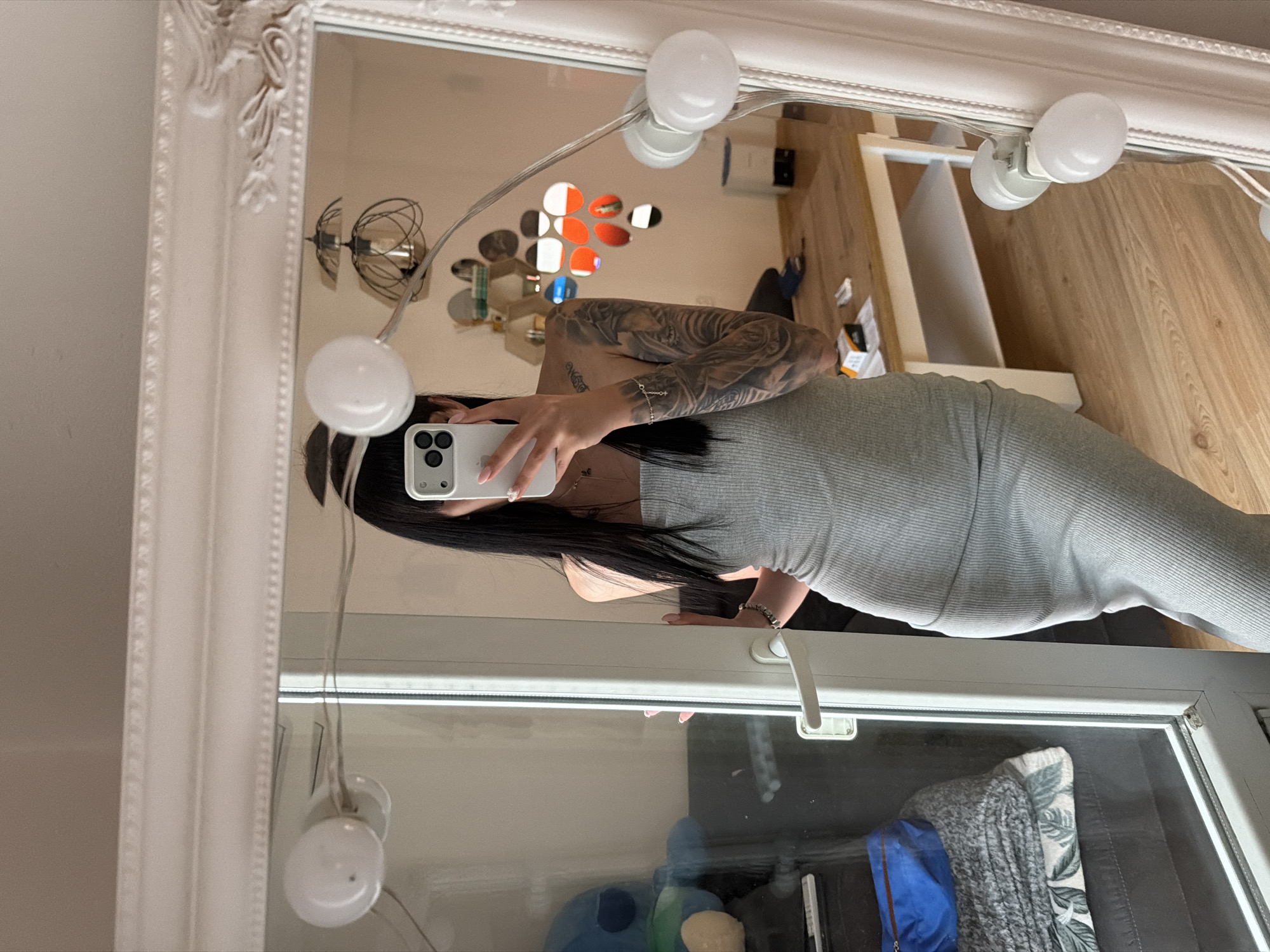A person is taking a mirror selfie in a room. The person has long black hair and is wearing a grey strapless dress. They have extensive tattoos on their left arm and shoulder. The mirror is large and ornate with a white frame. It is surrounded by multiple light bulbs, possibly for makeup application. In the reflection of the mirror, a wooden table with some items on it, a lamp, and a wall with decorative circular mirrors are visible. To the left of the mirror, a sliding glass door reveals a glimpse of another room with a blue stuffed animal and folded blankets. The flooring appears to be light-colored wood. The overall impression is that of a personal space, likely a bedroom or dressing area. The image was taken in Plovdiv, Bulgaria.