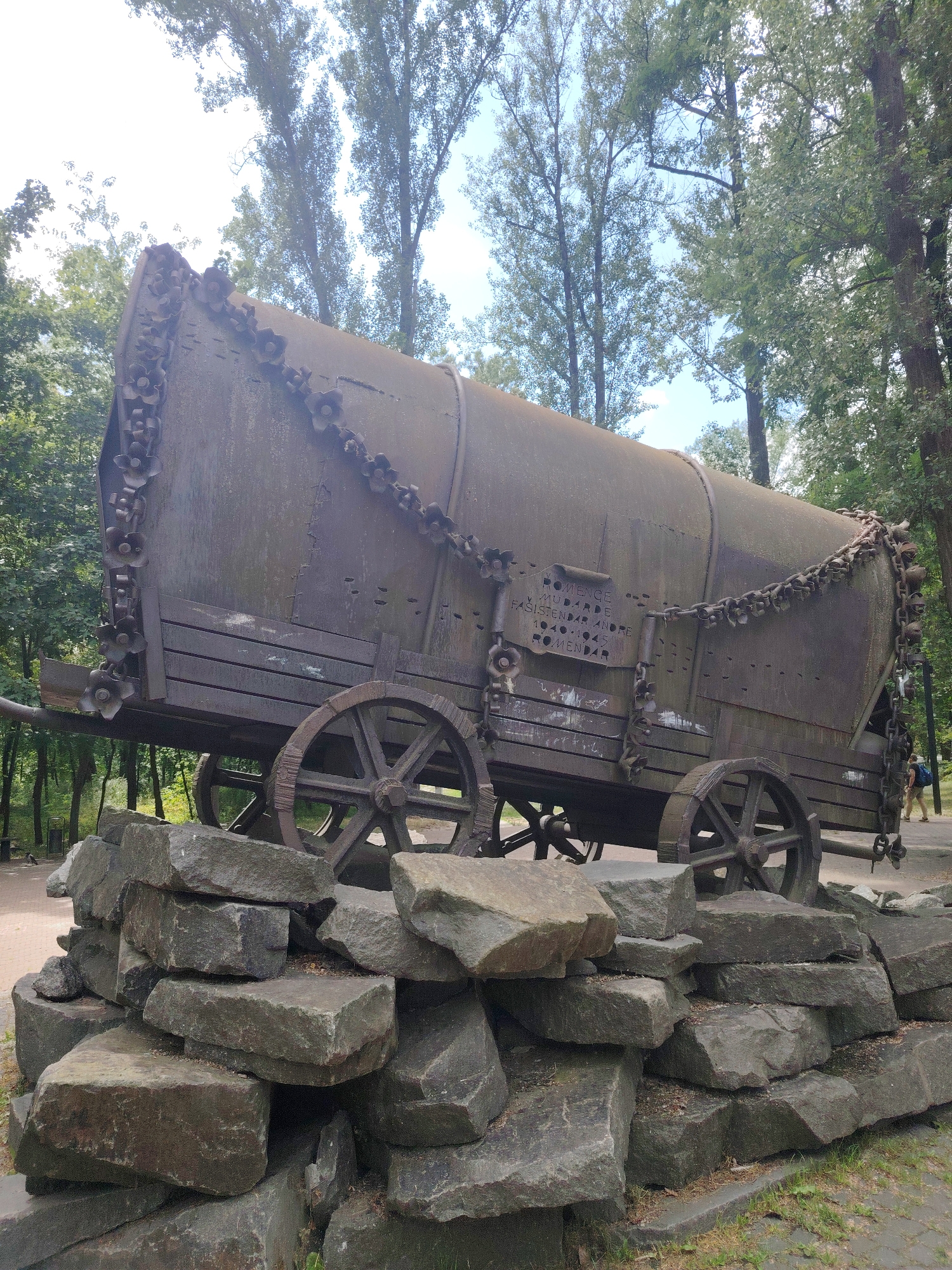 The image shows a monument made of a large, weathered, metal wagon with wooden planks and chains. It is positioned on a mound of large, rough-hewn stones. The wagon is decorated with metal flower-like elements along its edges and a prominent inscription is visible on its side. The monument is set outdoors in a park-like environment with tall trees and a pathway in the background. A person with a blue backpack is walking away from the monument in the background to the right. The monument is located in Babyn Yar park in Kyiv, Ukraine.