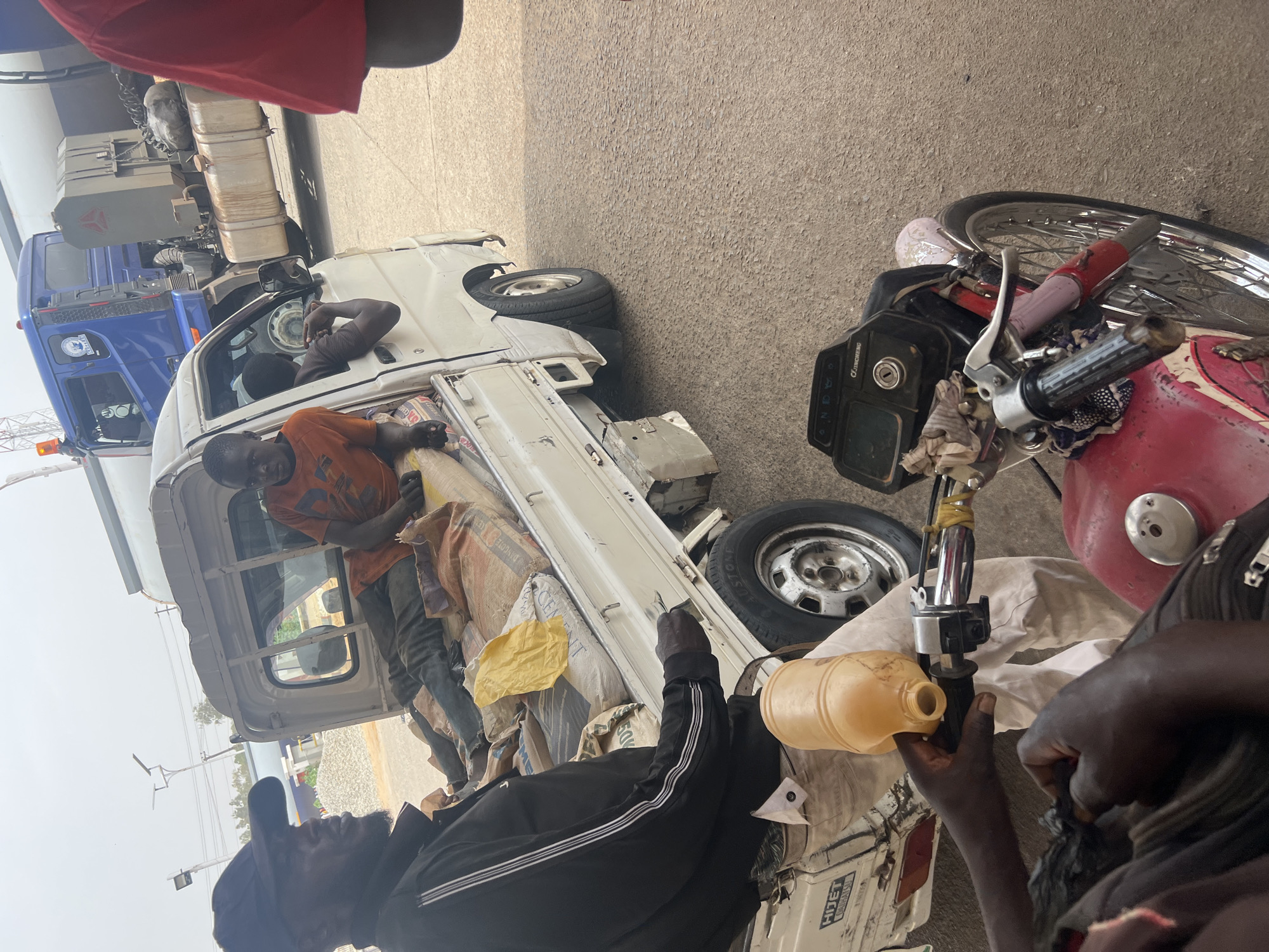 The scene shows what appears to be a roadside interaction in Jos, Nigeria. A white pickup truck is parked; it is loaded with sacks that appear to be cement. Two men are interacting with the truck, with one standing in the back and another on the ground close to a motorcycle. The man on the ground is filling the motorcycle's fuel tank with fuel from a yellow plastic container. The environment appears to be an open area with paved surfaces. A large blue and white truck is visible in the background.