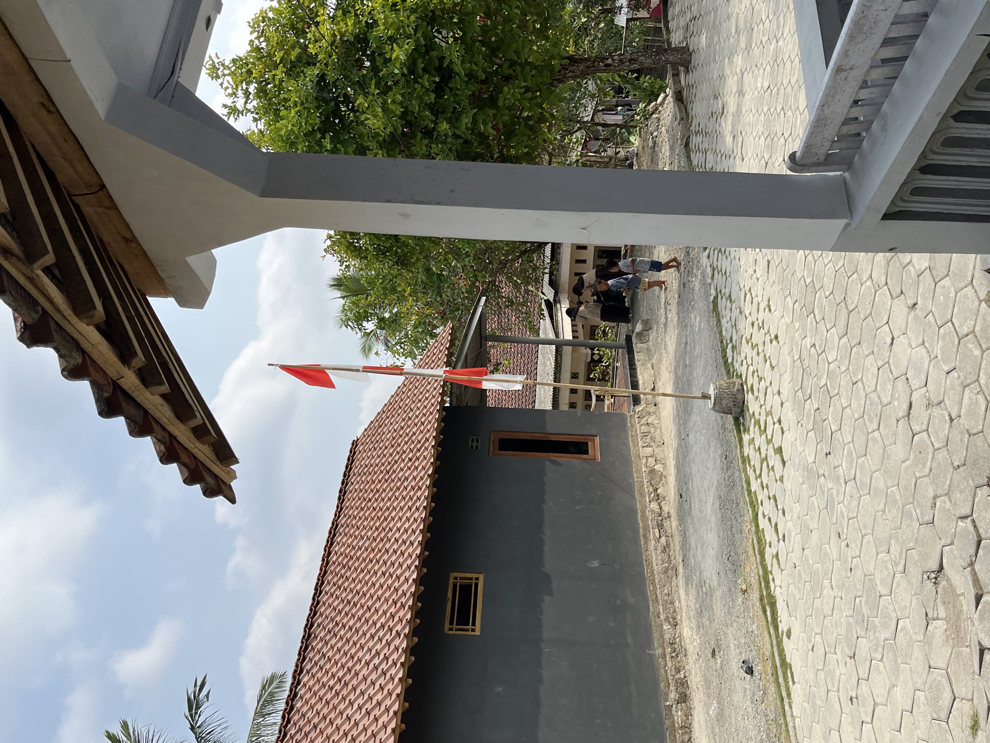 This is an outdoor, eye-level shot of a street scene in Kemangguhan, Indonesia. The weather appears to be sunny and clear, with a light blue sky and some scattered white clouds.
In the foreground, a portion of a grey, modern-looking porch railing is visible on the right. Beyond that, a cobblestone pathway extends into the scene.
To the left of the pathway, a dark grey building with a red tile roof is prominent. It has a small, horizontally oriented window with a wooden frame and a taller, vertical window with a wooden frame. In front of this building, a small, weathered concrete base supports a pole from which a red and white Indonesian flag is flying. The flag appears to be attached with tape and is flapping slightly.
Further into the background, a lighter-colored building with a similar red tile roof is visible. Several people are standing in front of this building, including two individuals wearing dark headscarves and two young children. One child is wearing a blue shirt and shorts, and the other is wearing a light-colored top and shorts. They appear to be interacting.
To the right of the people, a large, leafy green tree is in full bloom, its branches reaching towards the sky. Behind the tree, other structures and greenery are visible, suggesting a residential or semi-rural environment. A palm tree is also visible on the far left of the image. A pigeon is on the ground in the mid-ground.
