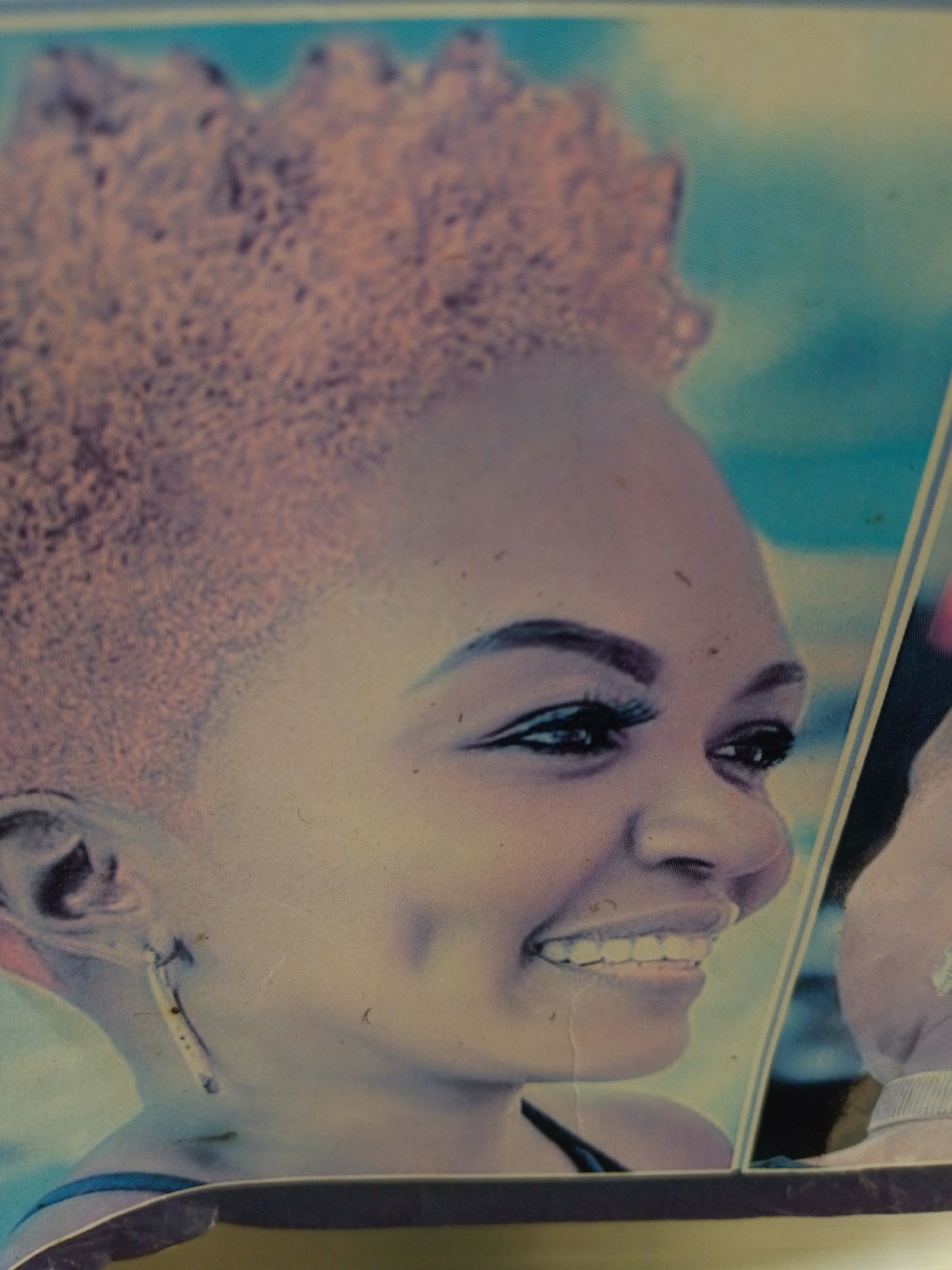 The image shows a close-up portrait of a young woman with short, curly, reddish-brown hair. She is looking to her right, with her head tilted slightly. Her left eye is visible, and she has a dimple on her left cheek. Her mouth is open, revealing her teeth in a smile. She is wearing an earring in her left ear, which appears to be a long, thin piece of metal with a circular detail at the bottom. The background is a blurred gradient of blues and purples, suggesting an outdoor setting, possibly a beach or sky. The overall image has a grainy texture and appears to be a photograph. The location in the city of Unknown, Unknown is not discernible from the image.