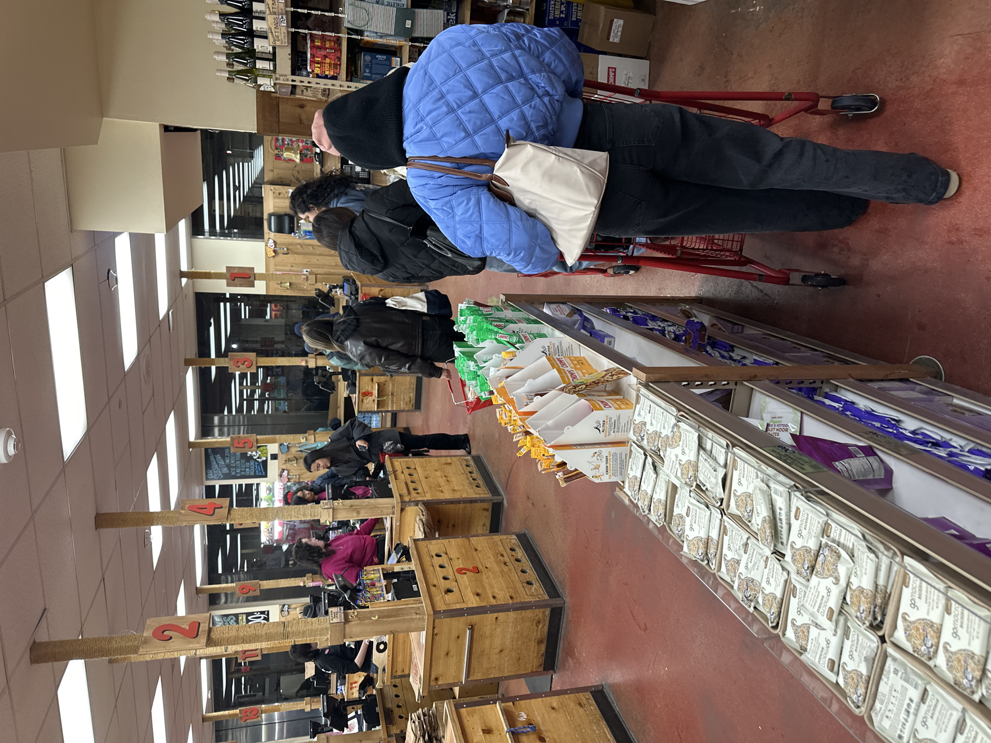 The image shows a queue of customers in a Trader Joe's grocery store. The location is most likely in New York City, USA, given the store brand is a popular nationwide chain. Several people stand in line at the checkout counters, numbered from 1 to 5. The counters are constructed with light-colored wood panels, and each has a tall post with a number sign. The customers are bundled in jackets and hats. The aisles feature grocery products like energy bars and dog treats. Shopping carts are visible. The floor is predominantly a terra-cotta red color. The lighting in the store is bright, suggesting fluorescent or LED fixtures. The image suggests a busy shopping environment.
