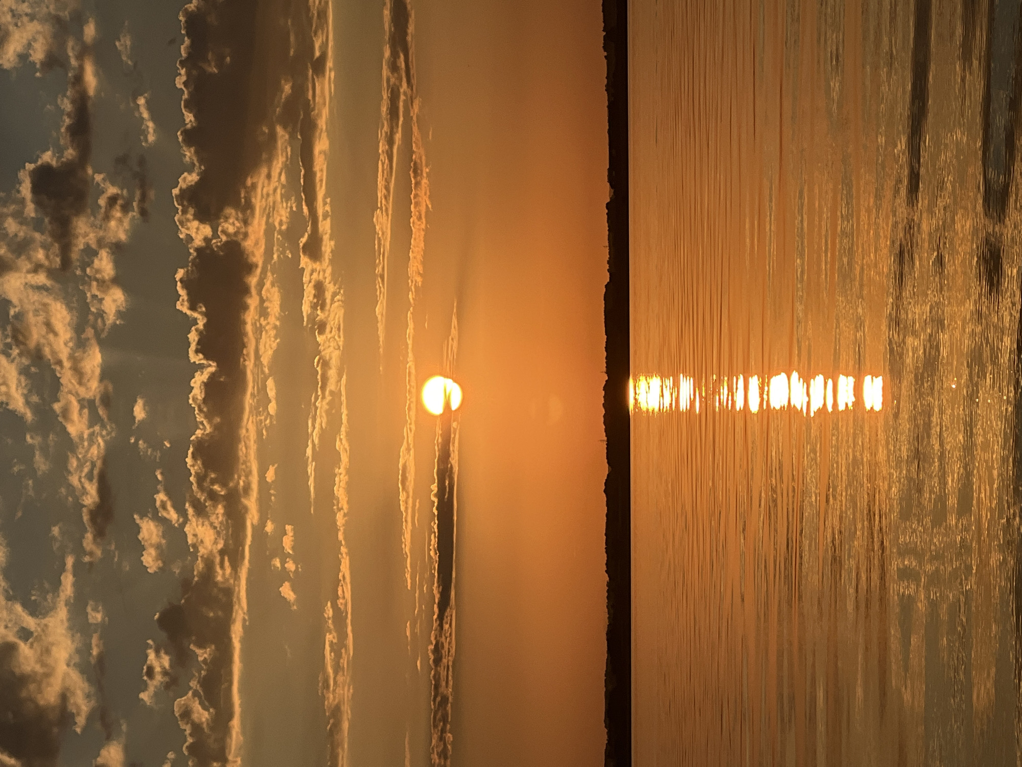 A sunset over a body of water is depicted in the image. The sky is filled with clouds that are tinged with orange and yellow from the setting sun. The sun itself is a bright orb, partially obscured by clouds. A reflection of the sun shimmers on the surface of the water, creating a golden path across the calm expanse. The horizon is lined with a dark silhouette of trees and land. The scene appears to be a serene natural landscape, likely in Novopichugovo, Russia, given the context of the prompt. There are no people or man-made structures clearly visible in the foreground or middle ground.