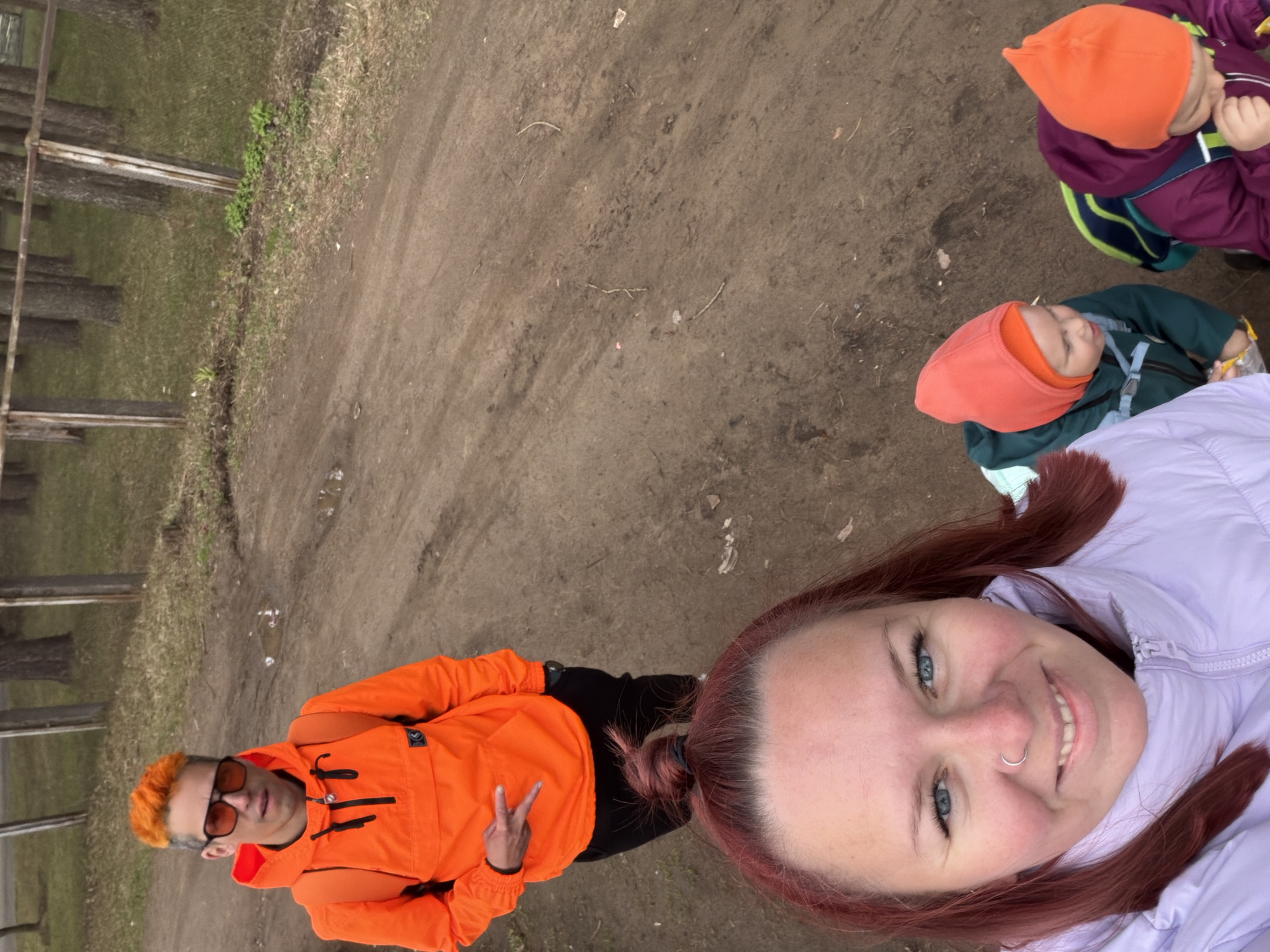 This sideways-oriented image captures a group of four people, two adults and two children, outdoors on a dirt path in Chaikovsky, Russia.
In the foreground, a woman with auburn hair, blue eyes, and a nose ring is taking a selfie. She wears a light purple jacket and smiles broadly at the camera, conveying a cheerful mood. Behind her, to the left, a man with vibrant orange hair and sunglasses is visible. He wears a bright orange jacket, black pants, and a backpack, and makes a peace sign with his left hand.
Further back, on the right side of the frame, are two young children. Both are wearing orange beanies; one is in a dark green jacket, and the other in a purple jacket with a green backpack. They appear to be looking down or away from the camera.
The setting is an unpaved, brownish dirt path, suggesting a rural or park environment. To the left of the path, there are patches of sparse grass and several bare trees, with what appears to be a simple wooden fence or railing running horizontally in the far background. The lighting is soft and even, indicating daytime, possibly under an overcast sky. The overall scene suggests a casual and pleasant family outing. No visible text is discernible in the image.
