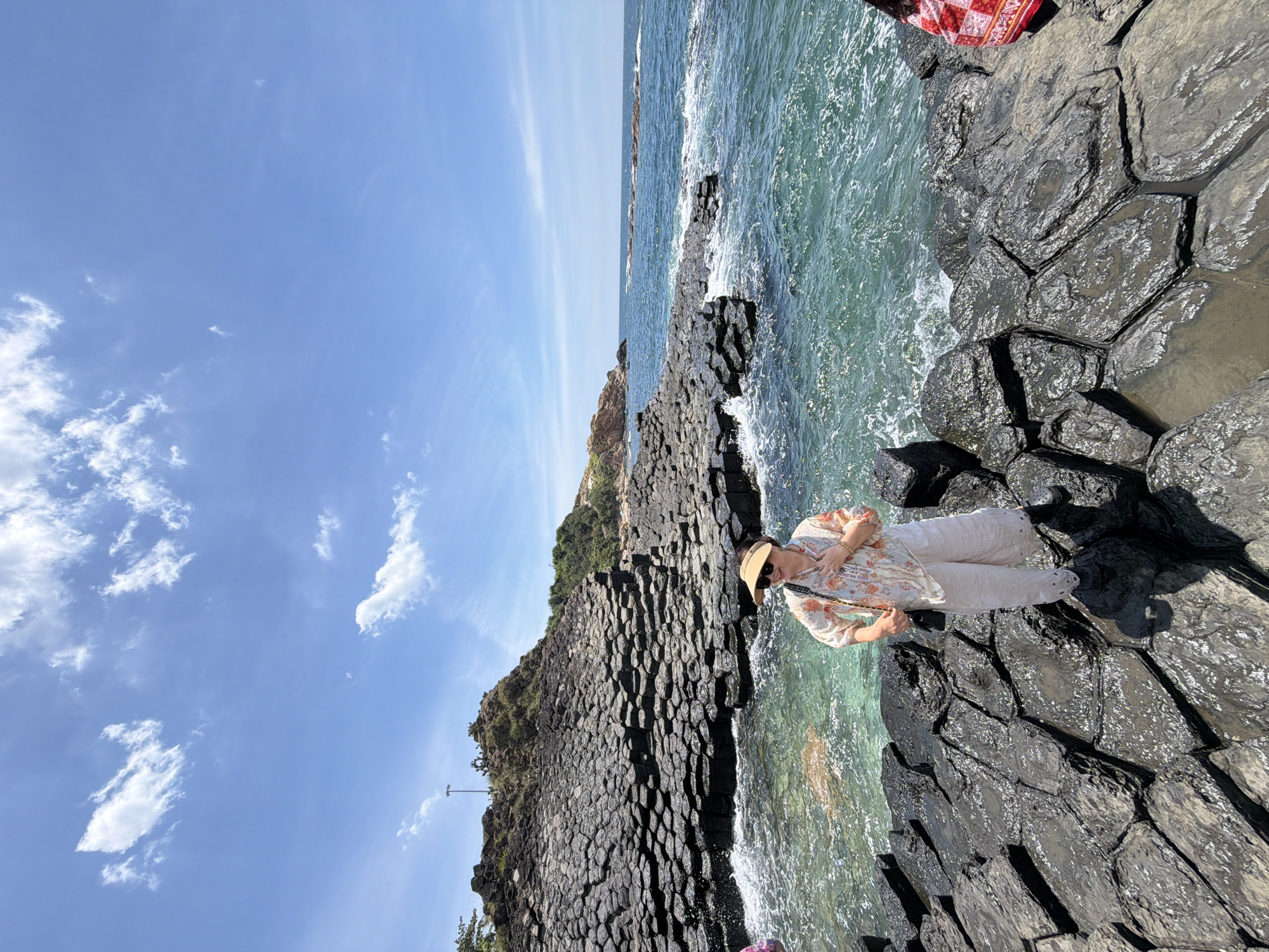 In a sun-drenched outdoor scene, a person stands on a unique geological formation of dark, hexagonal basalt columns near the ocean. The sky above is a clear, bright blue with scattered white clouds. The turquoise water laps against the rocks, creating small waves and white foam.
The person, facing away from the viewer and looking towards the water, wears a light-colored floral shirt and white pants, accessorized with a straw hat and sunglasses. They are holding a small purse with a chain strap. The hexagonal basalt columns, a notable geological feature, are wet and shiny, reflecting the sunlight.
The overall atmosphere suggests a pleasant day for sightseeing and enjoying the natural scenery. The location appears to be a coastal area with volcanic rock formations. The time of day is likely midday due to the bright sunlight and clear sky. No visible text is present in the image.