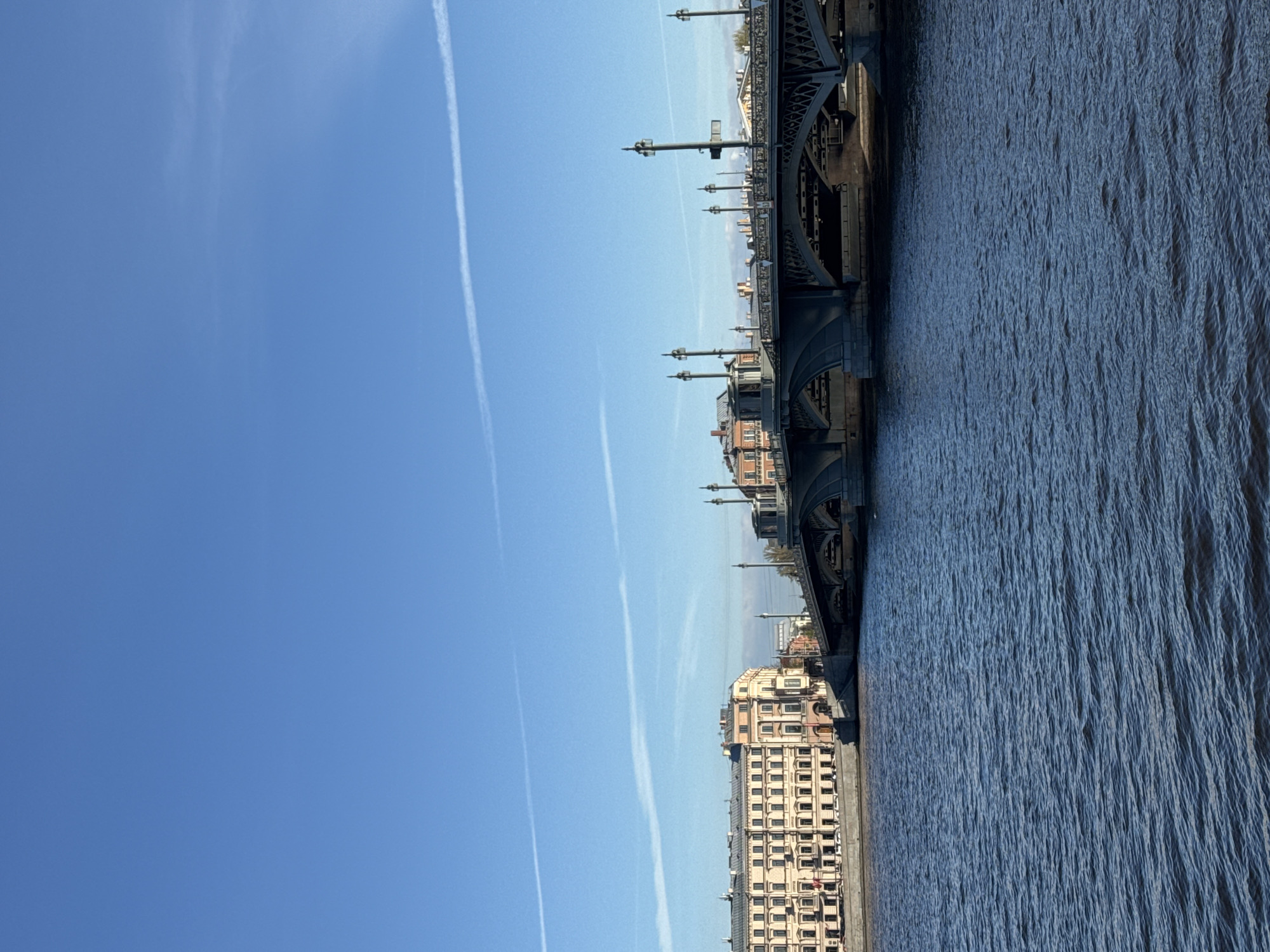 The image, when correctly oriented, presents a wide view of a cityscape dominated by a large body of water, a prominent bridge, and buildings under a clear sky.
**Subjects and Scene:** A broad expanse of dark blue water, featuring textured ripples on its surface, occupies the left side of the frame. To the right, a substantial, dark-colored bridge, likely constructed from metal or stone, spans the water. This bridge showcases intricate structural details, including visible arches and a series of decorative lamp posts mounted along its upper span. On the far bank, situated beneath the bridge and extending further into the left background, are several multi-story buildings. These structures have light-colored facades and numerous windows, reflecting a classical or historic European architectural style, consistent with an urban environment. The upper portion of the image is filled by a bright, clear blue sky.
**Notable Details:** The sky is marked by several thin, white contrails left by aircraft, streaking horizontally across the blue expanse. The scene is illuminated by natural light, indicating it is daytime with clear weather. There are no people or specific activities prominently visible, contributing to a serene and expansive atmosphere. No discernible text is present in the image. The architectural characteristics of the bridge and buildings, combined with the presence of a wide waterway, align well with the provided context of Saint Petersburg, Russia.