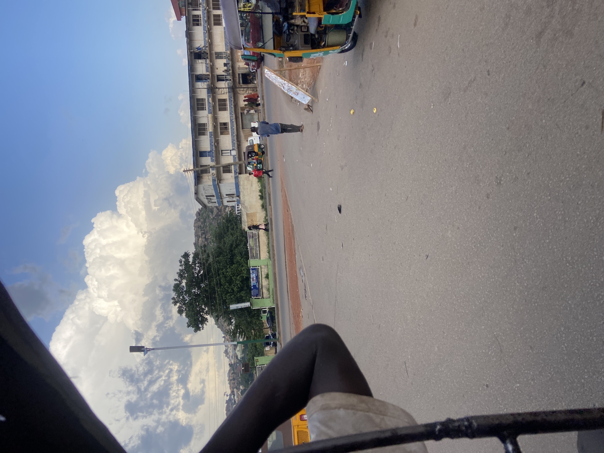 The image depicts a street scene in Jos, Nigeria, likely during the daytime. The perspective is from inside a moving vehicle, possibly a bus or car, with part of the vehicle's interior visible in the foreground. The road surface is asphalt, and there are several vehicles on the street, including a colorful auto-rickshaw and a white van.
Several people are visible, some walking on the side of the road and others inside vehicles. One man in a blue shirt and dark pants stands in the middle of the road, facing away from the camera. Another person in a red shirt is walking.
A multi-story building with a white facade and blue trim lines the left side of the street. The building has numerous windows and balconies. In the background, there are hills covered with trees and rocks, suggesting a somewhat hilly or mountainous terrain. The sky is partly cloudy with large, white cumulus clouds dominating the upper left portion of the image.
The scene appears to be a typical urban street with a mix of buildings, vehicles, and pedestrian activity. The overall impression is one of a busy, sunny day in a Nigerian city. No discernible text is visible in the image. The orientation of the image is sideways, with the sky appearing on the left and the ground on the right.