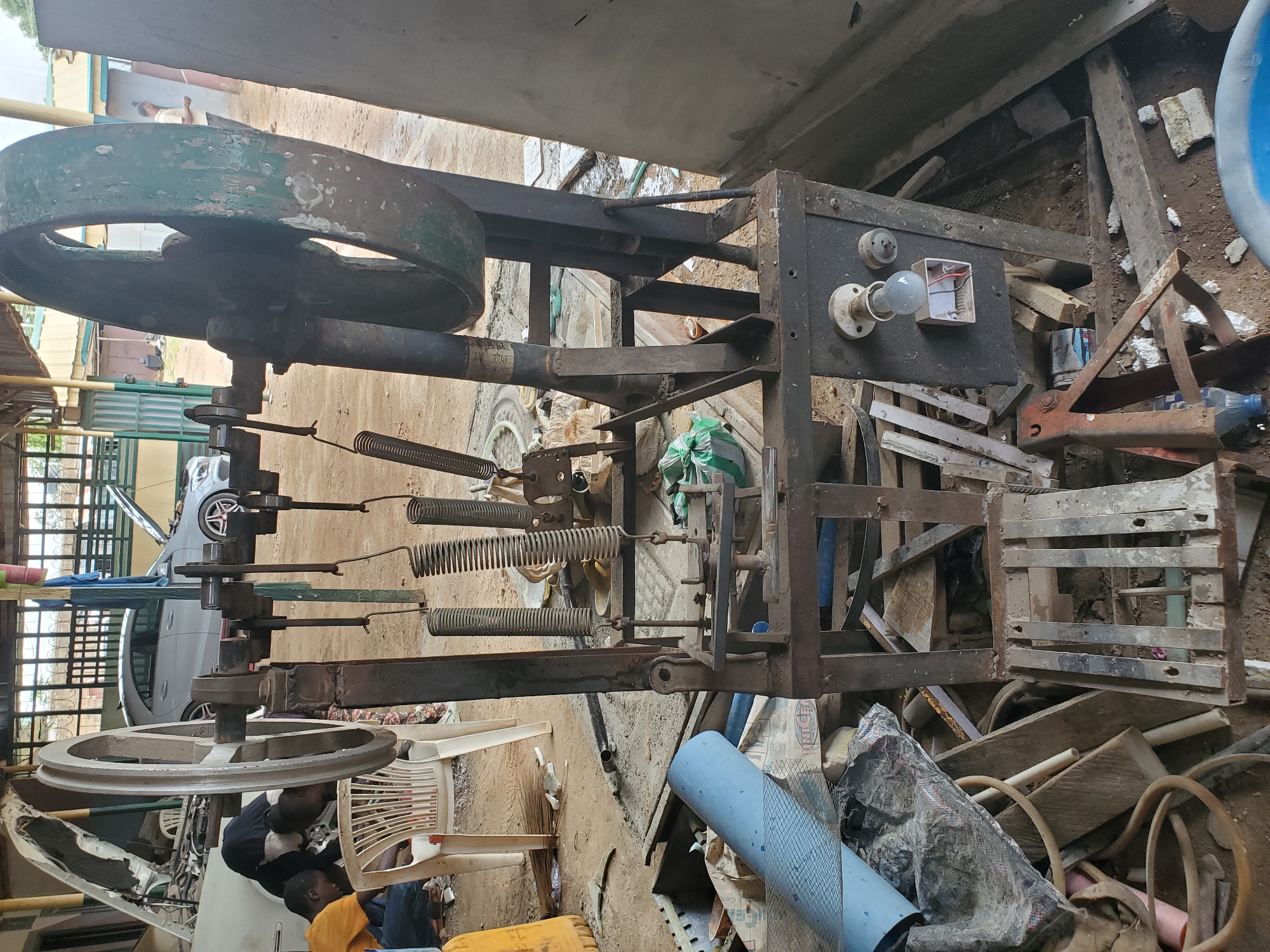 The image depicts an outdoor workshop scene, likely in Jos, Nigeria, with a focus on an old, heavy-duty machine. The machine is predominantly metal, with a large flywheel at the top, painted green but showing significant wear and chipping. Below the flywheel, a complex assembly of springs, levers, and metal components suggests a functional purpose, possibly for stamping or pressing. A lightbulb is mounted on a metal panel at the base of the machine, along with a socket and some exposed wiring, indicating it may be powered.
In the background, two individuals are seen hunched over, appearing to work on a white car with its hood open. A light-colored sedan is also visible to the left, also with its hood up. The environment is cluttered with various materials, including pipes, bags, wooden planks, and what appears to be part of a blue plastic barrel or container. The ground is a mix of dirt and some concrete or paved areas. The overall impression is one of a busy, utilitarian space where mechanical repairs or fabrication take place.