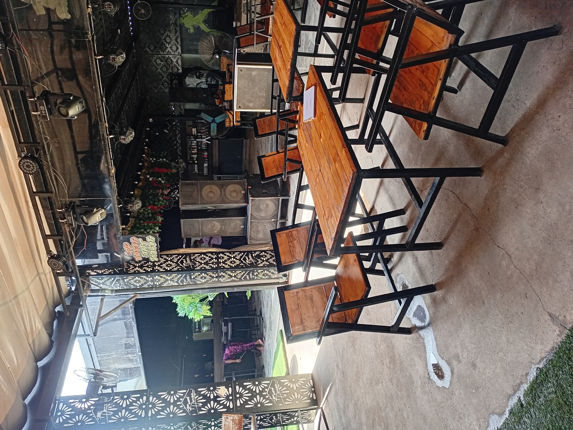 The image shows the interior of an outdoor bar or restaurant in Howayi, Nigeria. The scene is furnished with wooden tables and chairs, some of which are empty, while others are occupied by customers. A person in patterned leggings is bent over at a bar in the background, and another person stands near a set of speakers. The space is decorated with decorative panels featuring floral motifs, and a sign that reads "STOP THINKING START DRINKING" is visible. Overhead, lighting equipment and speakers are suspended from a metal frame. The floor is concrete, with some wet patches and cracks, and a section of artificial grass is present in the foreground. The overall atmosphere suggests a lively, yet partially empty, establishment.