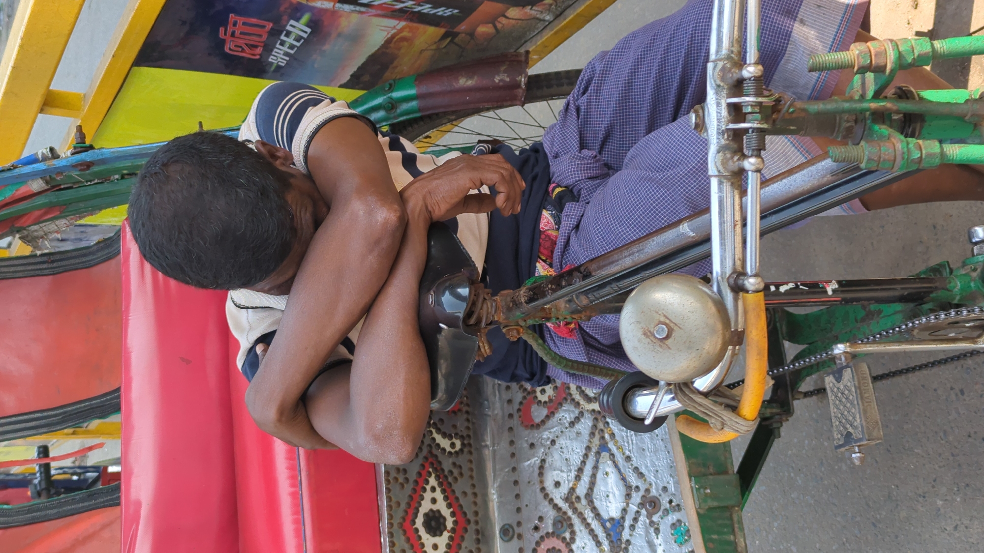 In Dhaka, Bangladesh, a man with dark hair rests with his arms crossed, his head down. He is wearing a striped shirt and is seated on a red cushion in a rickshaw. The rickshaw features a metal frame and patterned metal floor, and the vehicle has a bell, bicycle chain, and yellow and green paint.