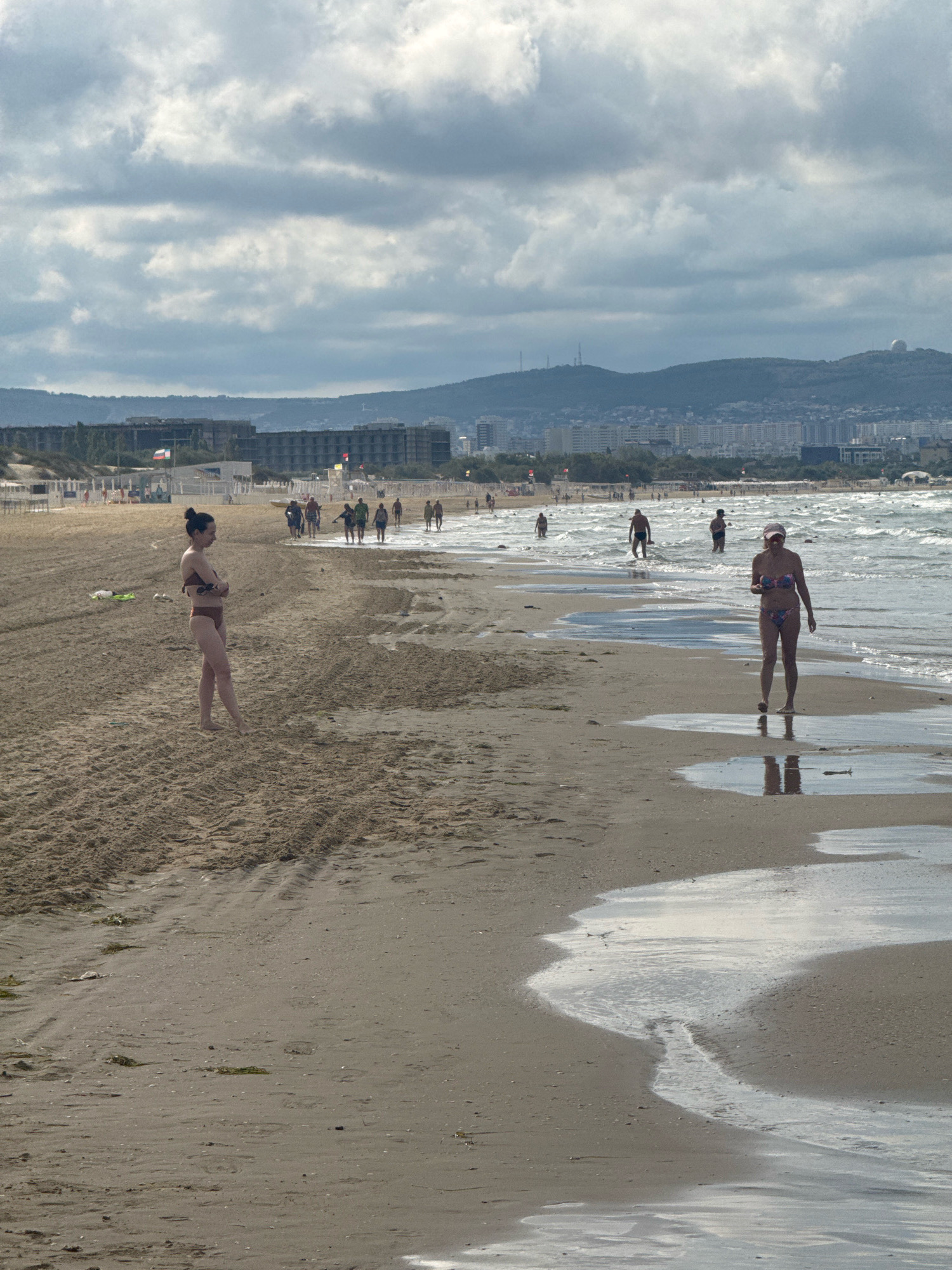 The image depicts a beach in Anapa, Russia, with a wide sandy expanse stretching towards a choppy sea under a cloudy sky. In the foreground, a woman in a brown bikini stands with her arms crossed, facing away from the camera. Further down the beach, closer to the water, another woman in a patterned bikini walks towards the viewer, holding an object in her hand. Several other individuals are visible in the distance, some wading in the shallow water and others walking along the shoreline. The background reveals a cityscape with numerous buildings and hills, suggesting a populated coastal area. The sand shows tire tracks and some scattered debris. The overall atmosphere is that of a moderately busy beach on an overcast day.