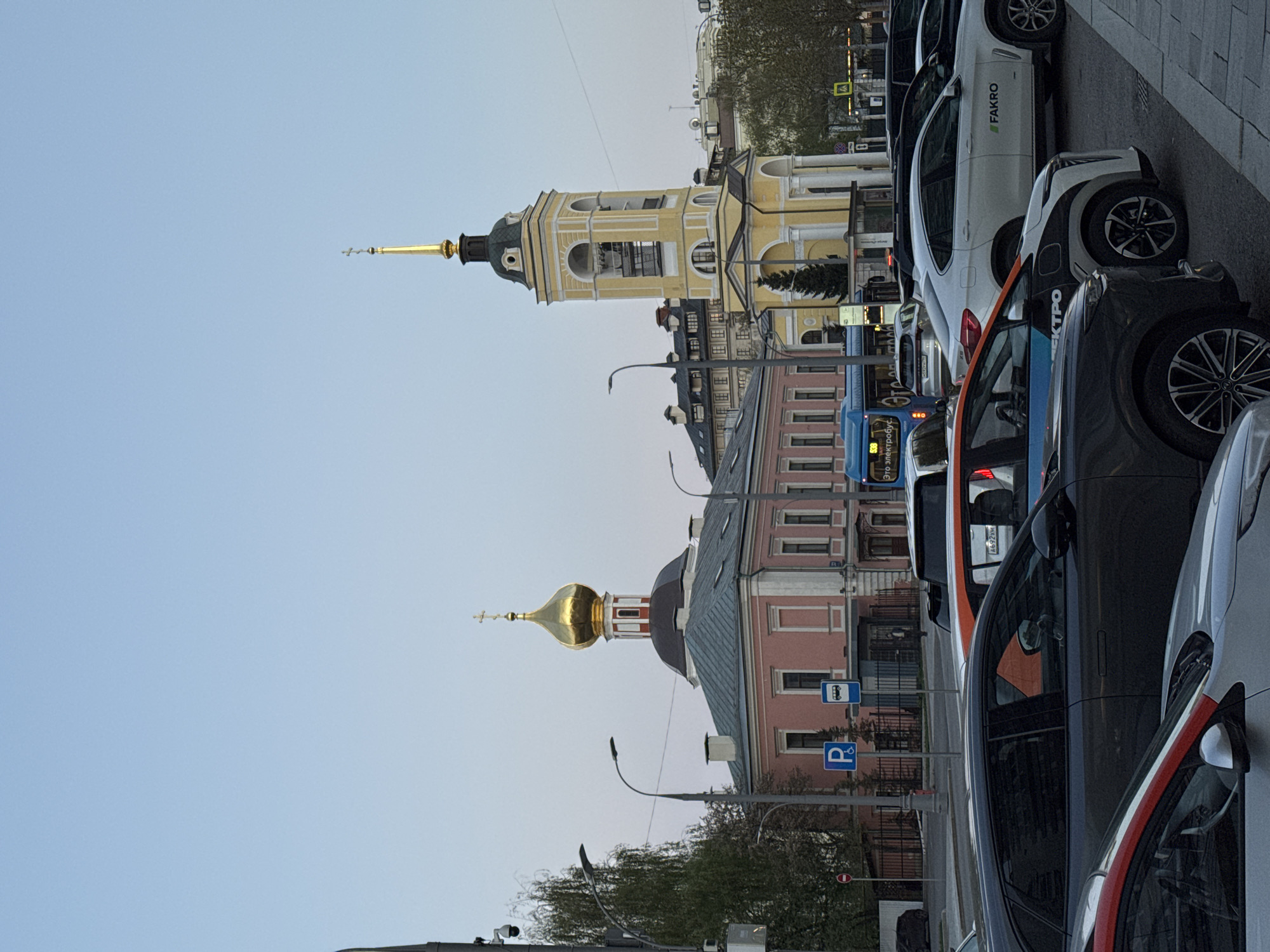 The image depicts a street scene in Moscow, Russia, showcasing architecture and urban elements. Dominating the view are two churches with prominent spires. One has a gold dome and cross, while the other features a taller yellow and white bell tower topped with a spire. Several buildings are visible in the background, including a pink building with a gabled roof. The street features multiple parked cars on the right side of the frame, including vehicles with corporate branding. Street signs, including those for parking and public transportation (bus stop), are visible. A blue bus is traveling on the road. Overhead power lines run parallel to the street. The sky is a light blue, indicating an overcast day.
