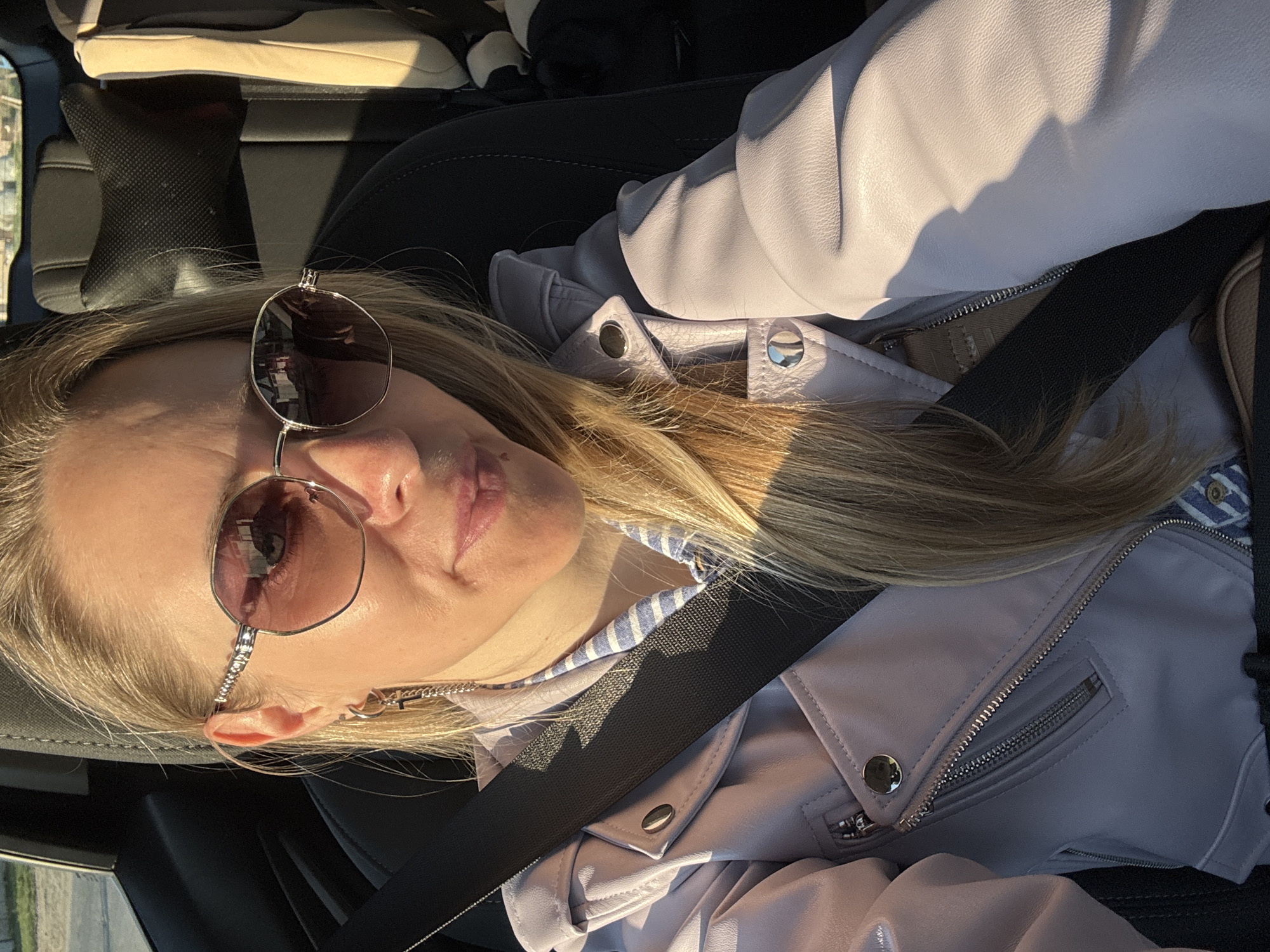 A blonde woman wearing sunglasses and a lavender leather jacket is seated in a car, wearing a seatbelt. Her hair is blonde and long. The car's interior is visible. The location is unknown but the description includes an instruction about Chaikovsky, Russia.