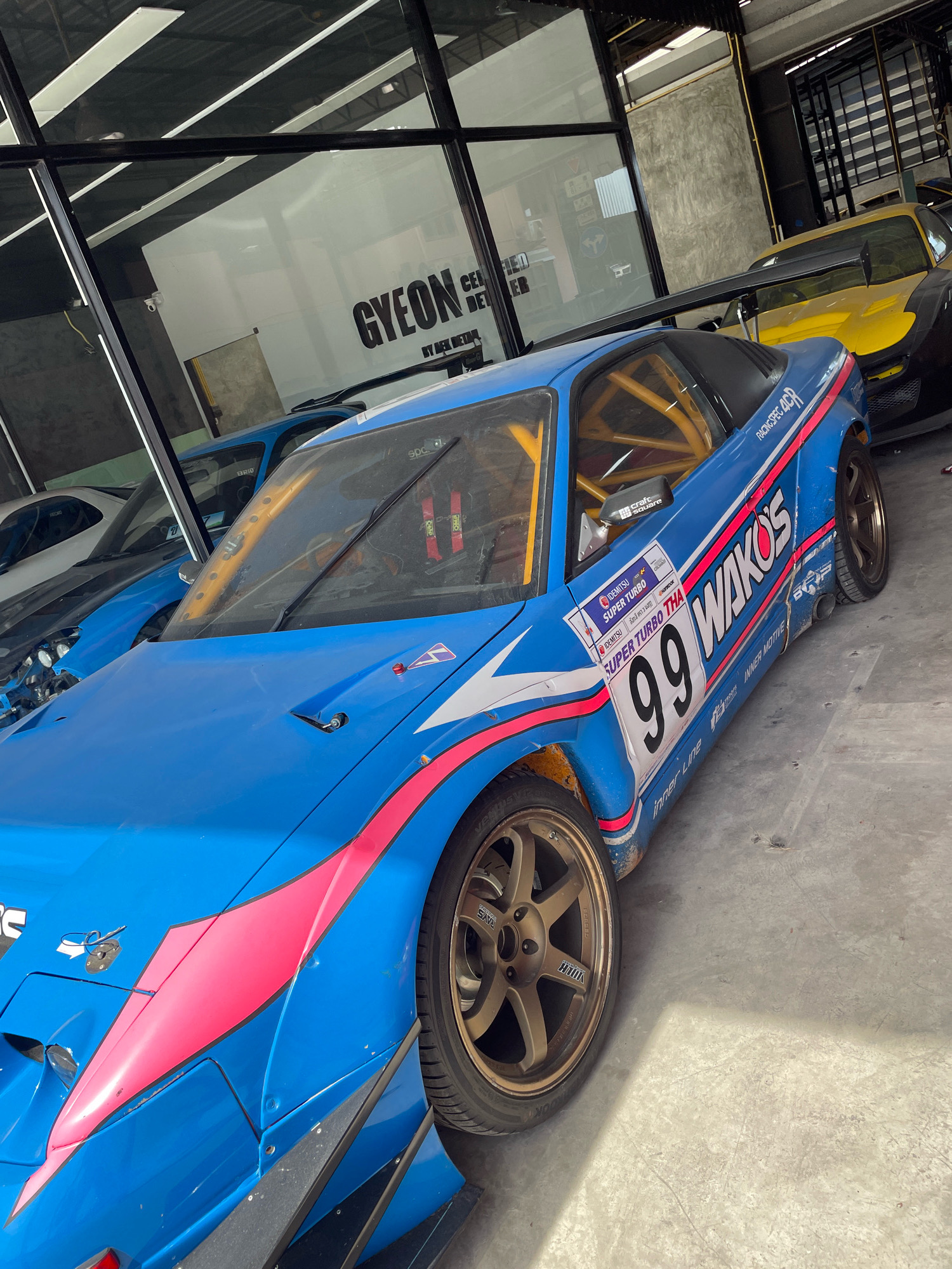 The image depicts a blue sports car with racing decals, including the number 99, parked in what appears to be a garage or workshop. The car has a prominent spoiler and racing cage visible through the windows. A yellow sports car is parked behind it. The setting is indoors with a concrete floor and a metal roof structure. A large window with the text "GYEON CERTIFIED DETAILER BY BEN DĒTAILING" is visible on the left. The location is identified as Nong Prue, Thailand. There are no people visible in the image.