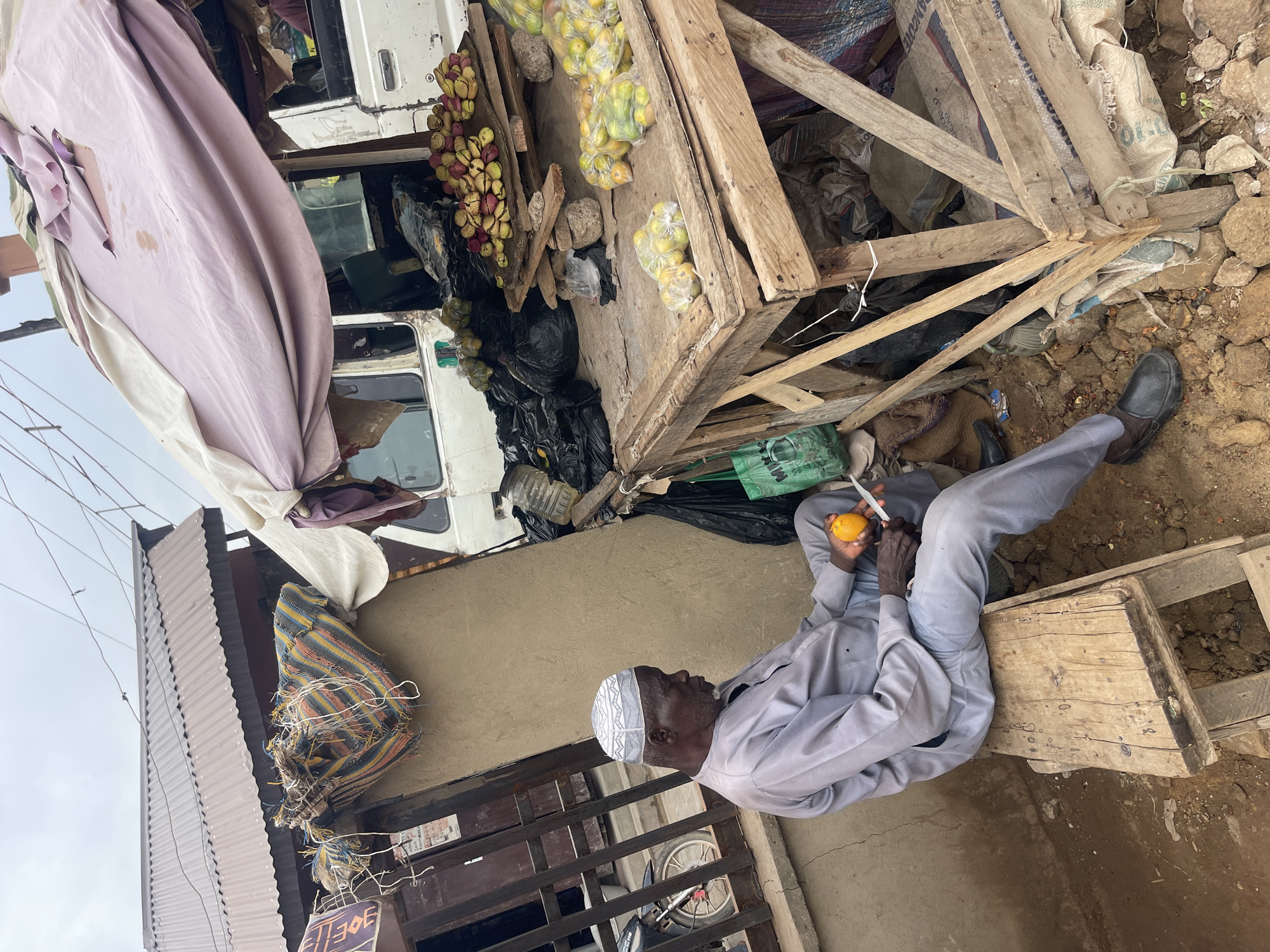 An elderly man in Zaria, Nigeria sits on a wooden stool outdoors, wearing a grey robe and a patterned white skullcap. He holds an orange in his left hand and a knife in his right, peeling the fruit. The man sits adjacent to a makeshift wooden structure displaying various fruits and vegetables, including apples, bananas, and oranges. A white truck is visible in the background, partially covered by a large, dirty purple tarp. Black trash bags and various containers are visible under the wooden structure. The area around the man is a mix of dirt and pavement. A fence and a building are behind him.