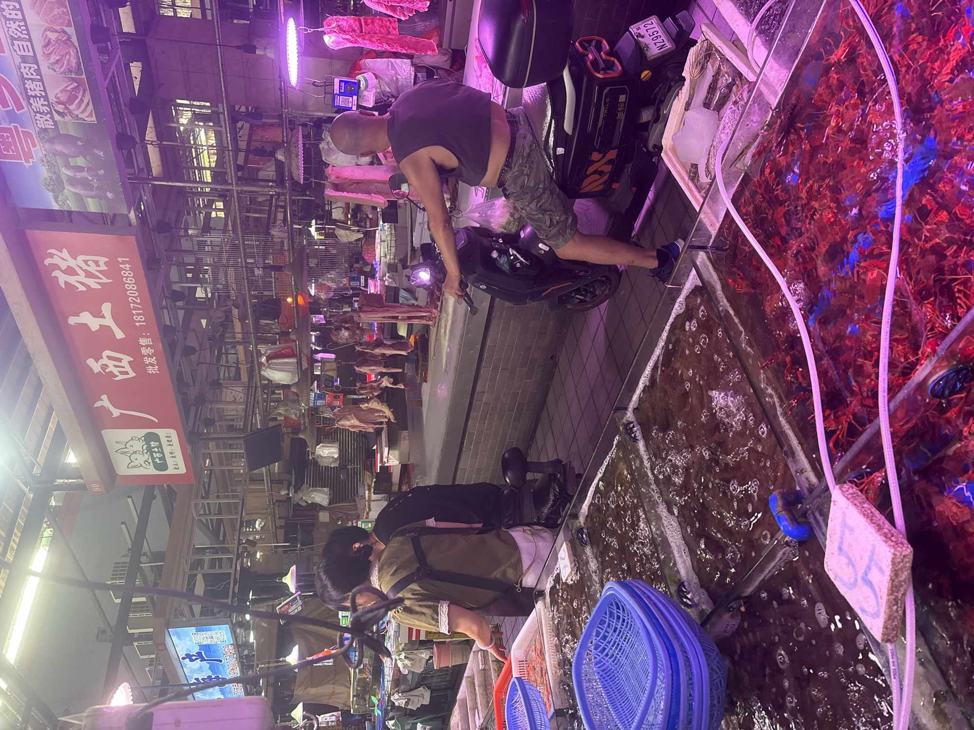 The image depicts a bustling market scene, likely in the city of Nanhai District, China, under artificial lighting that casts a purple hue. In the foreground, large glass tanks filled with water contain live seafood, primarily what appear to be lobsters or crayfish, with some blue elements visible in the water. Blue plastic baskets are stacked on the edge of one tank. Further back, a man with a shaved head, wearing a dark sleeveless shirt and camouflage shorts, is seated on an electric scooter. He is facing away from the camera, looking towards a display of meat products hanging from hooks above a counter. The meat section features signage in Chinese, including the characters "广西土猪" (Guangxi Native Pig) and images of pigs and pork cuts. Several people are visible in the background, some interacting with market stalls. The overall environment suggests a busy wholesale or retail market specializing in fresh produce and meat.