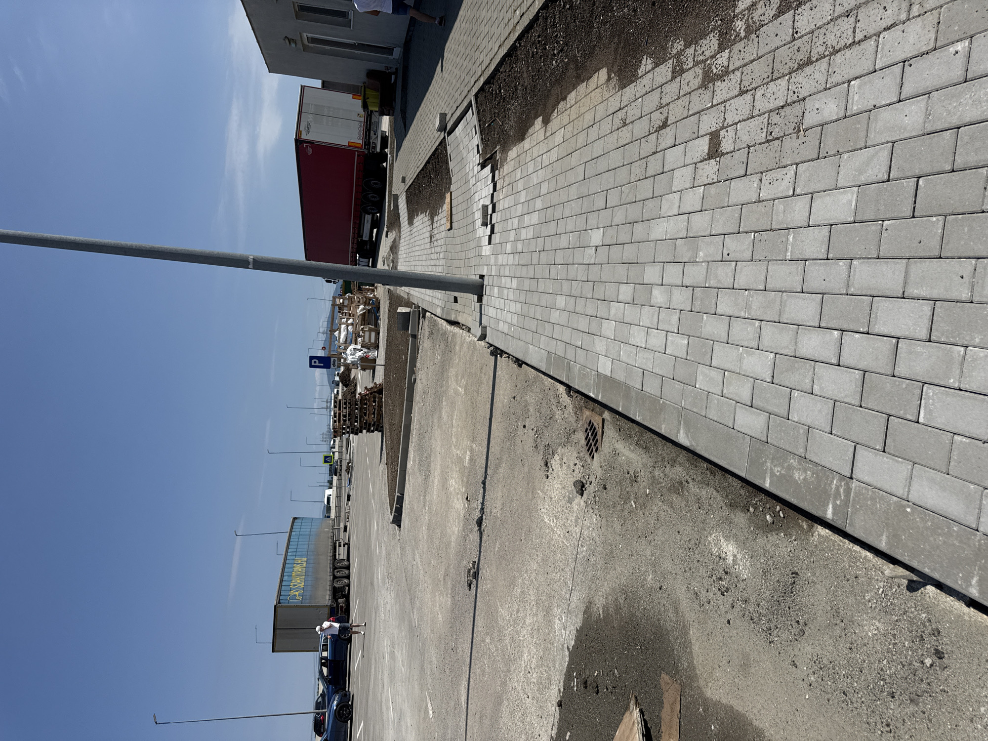 The image depicts an outdoor scene in Sanpetru, Romania, likely a commercial or industrial area. In the foreground, a newly paved pedestrian walkway made of light gray rectangular pavers runs diagonally across the frame, bordered by a dark brown dirt area. The walkway is incomplete in sections, with some pavers missing or misaligned. Behind the walkway, a grey concrete surface, possibly a parking lot or roadway, shows signs of wear and disrepair, with patches of dirt and gravel.
Further in the background, a large blue truck with the text "SZAKTRANS.HU" is parked. To its right, a red truck is also visible. Several wooden pallets are stacked near the red truck. A modern building with white walls and dark-framed windows is situated on the far right side of the image, partially obscuring the red truck. A tall grey pole, likely a lamppost, stands prominently in the mid-ground. The sky is clear and blue. A few individuals, dressed in casual clothing, are seen in the background, one near the blue truck and another near the building. The overall impression is one of ongoing construction or renovation in a functional outdoor space.