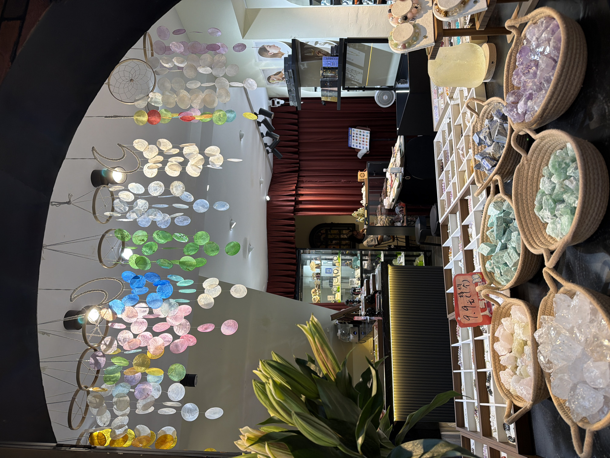 The image shows the interior of a retail store, potentially in the city of Chongqing High-tech Zone, China. The scene is observed from the store's entrance, where colorful circular mobiles hang from the ceiling. Further inside, a display case of jewelry is visible, and red curtains line the wall. To the right are shelves filled with crystals in baskets. In the foreground, a potted plant partially obscures the view.