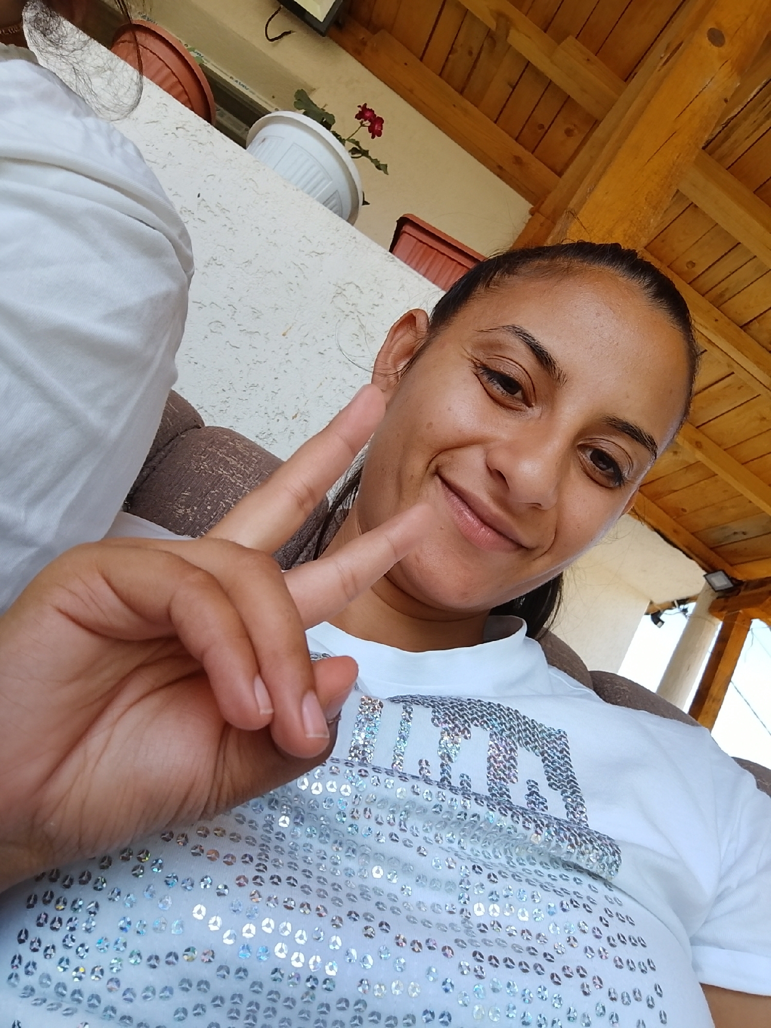 A woman is pictured in an outdoor, covered setting, possibly a patio or balcony. She is wearing a white t-shirt adorned with numerous silver sequins forming a pattern or text across the front. Her right hand is raised, displaying a peace sign. Behind her, the background includes textured walls, wooden beams of a roof structure, and hints of greenery from potted plants. The image's composition suggests it was taken by the woman herself, likely using a mobile device. The overall impression is casual and relaxed.