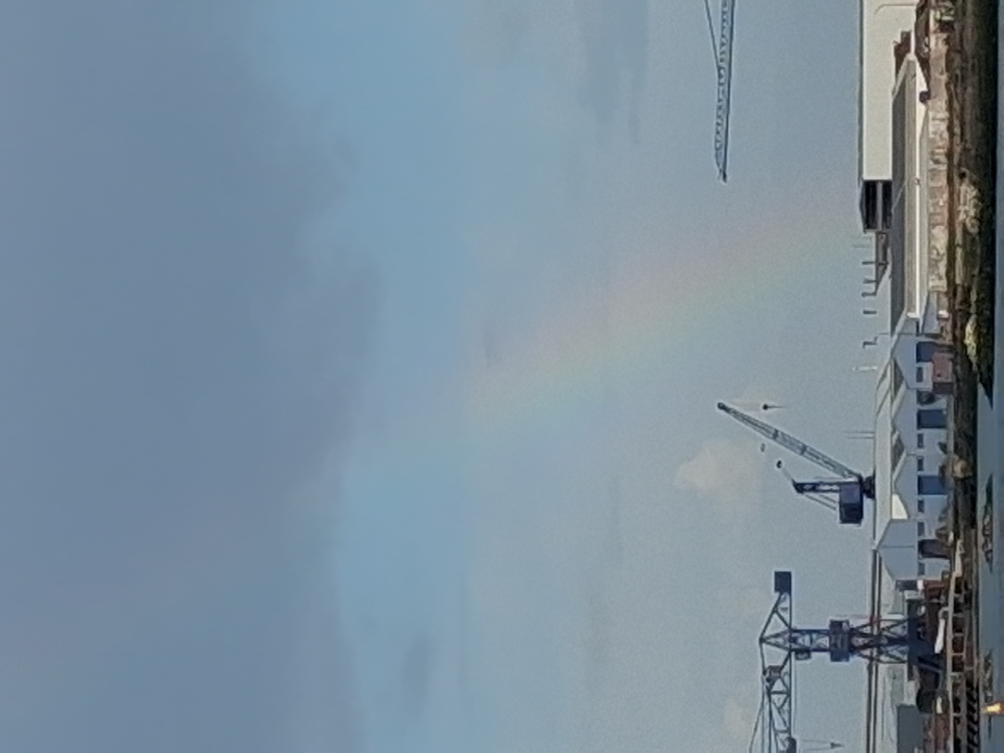The image presents a partially visible rainbow in the sky over what appears to be an industrial area in Figueira da Foz, Portugal. The environment features several cranes and buildings, possibly a port or shipyard, with the sky exhibiting a muted coloration due to the weather conditions associated with the rainbow formation. No people are visible in the shot. The picture is taken from a static, slightly elevated perspective.