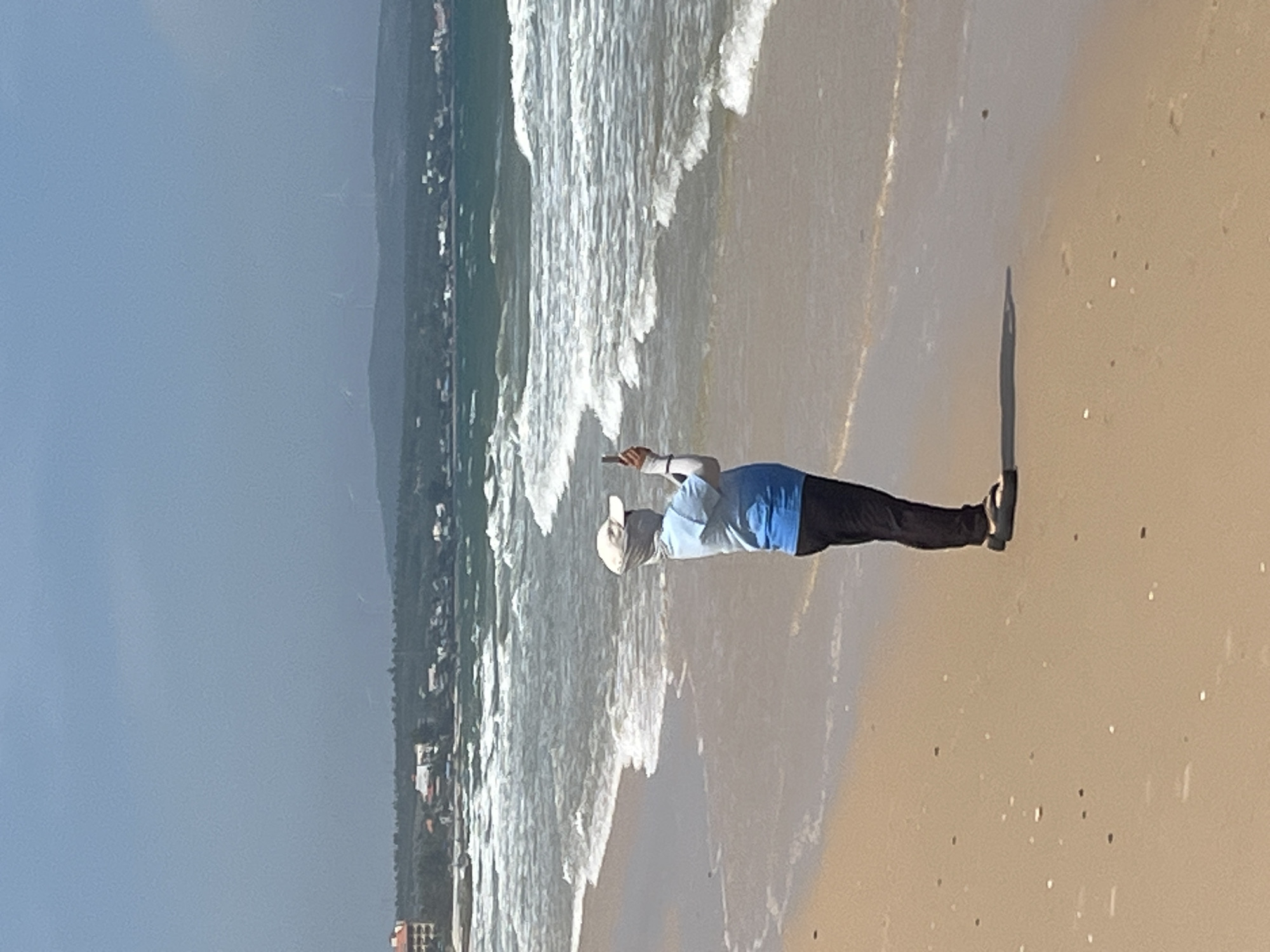 The image depicts a person standing on a sandy beach, with the ocean and waves in the background. The person is wearing a light blue shirt, dark pants, and a white cap. They are holding a dark object, possibly a phone, in their right hand, and appear to be taking a picture or recording a video. The waves are breaking on the shore, creating a white foamy pattern.
In the distance, a coastline with buildings and vegetation is visible. The sky is clear and blue, suggesting a sunny day. The lighting indicates it is daytime, likely mid-morning or afternoon. The sand is a light brown color, with small scattered pebbles or shells. The overall scene conveys a sense of leisure and enjoyment of the beach environment. The presence of buildings and the specific color of the sand and vegetation could be location cues, but without more context, it's difficult to pinpoint the exact location. The image is captured from a low angle, emphasizing the person and the beach. There is no visible text in the image.