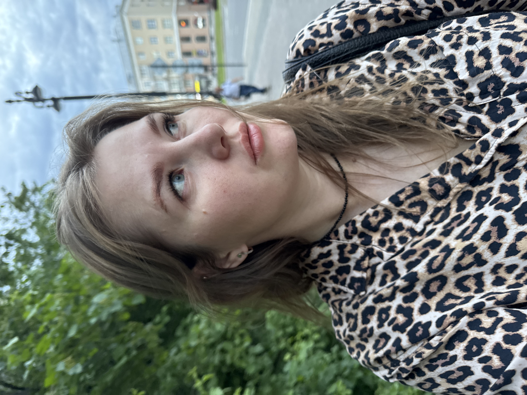 A woman with fair skin, blue eyes, and light brown hair poses in a medium shot taken from a low angle. She is wearing a leopard-print top and a thin, dark necklace. The background shows a street scene with a light-colored building, street lamps, trees, and a few pedestrians. The city is Perm, Russia.