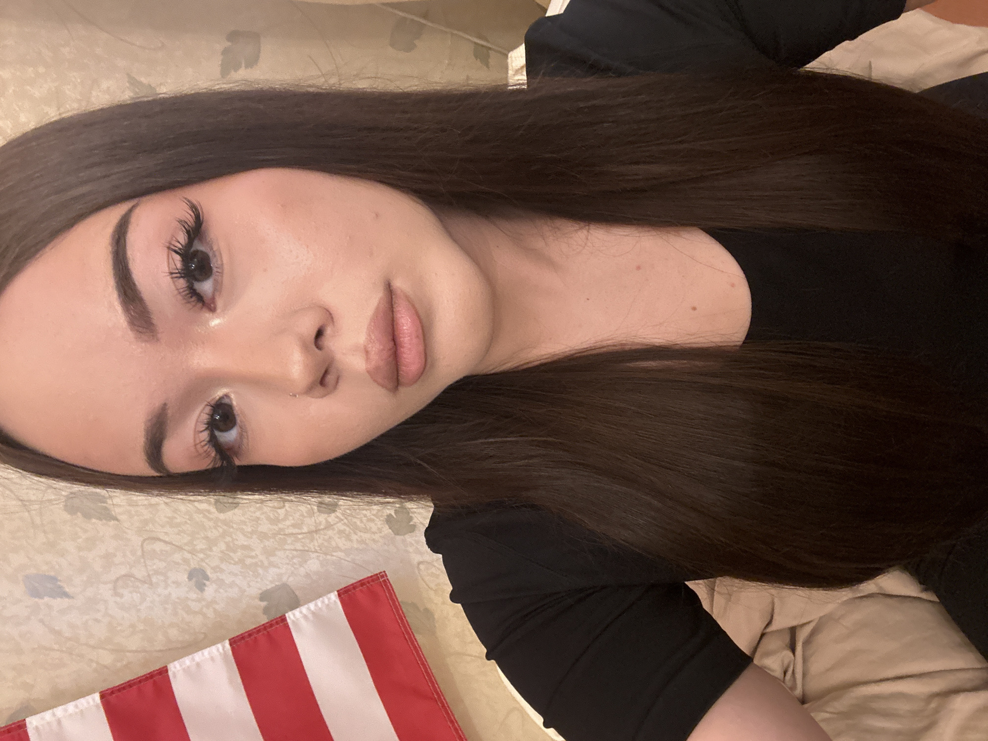 The image shows a close-up, slightly overhead portrait of a young woman reclining indoors. Her head is tilted, and she looks directly at the camera with a neutral and composed expression. She has long, straight, dark brown hair that cascades around her shoulders and over her body. Her face features clear skin, well-defined eyebrows, elaborate false eyelashes, and a natural-toned lip color. A small stud nose piercing is visible on her right nostril. She is wearing a black, long-sleeved top.
The background behind her head appears to be a light beige or off-white fabric with a subtle, possibly floral or leaf-like, pattern. To her right, a plain beige blanket or duvet is visible. In the bottom left corner of the frame, a small, red and white striped flag or fabric piece is partially visible, resting on the patterned surface. The lighting is soft and even, suggesting an indoor setting. The scene is set in Naberezhnye Chelny, Russia. There is no visible text in the image.