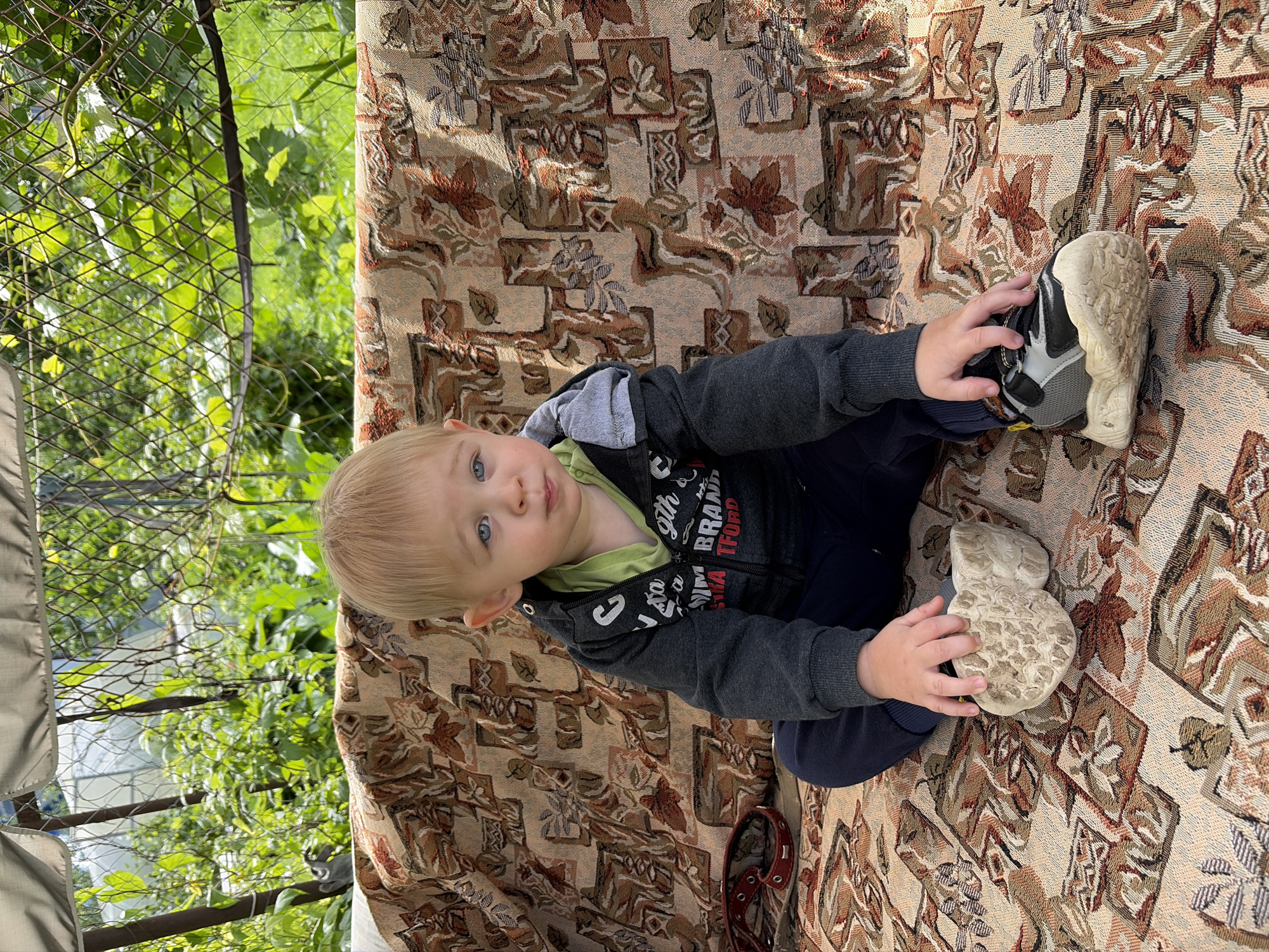 A young boy with blond hair and blue eyes sits on a patterned outdoor swing seat. He is wearing a gray zippered hoodie over a lime green shirt and dark blue pants. His hands are resting on his knees, and his shoes are visible: one appears to be a light-colored sneaker, and the other is a dark and gray athletic shoe. The swing is positioned outdoors, with a background of lush green foliage and a wire fence. A red collar is visible on the swing beside the boy. The scene is illuminated by sunlight, casting shadows on the patterned fabric of the swing. The location is specified as Kimry, Russia.