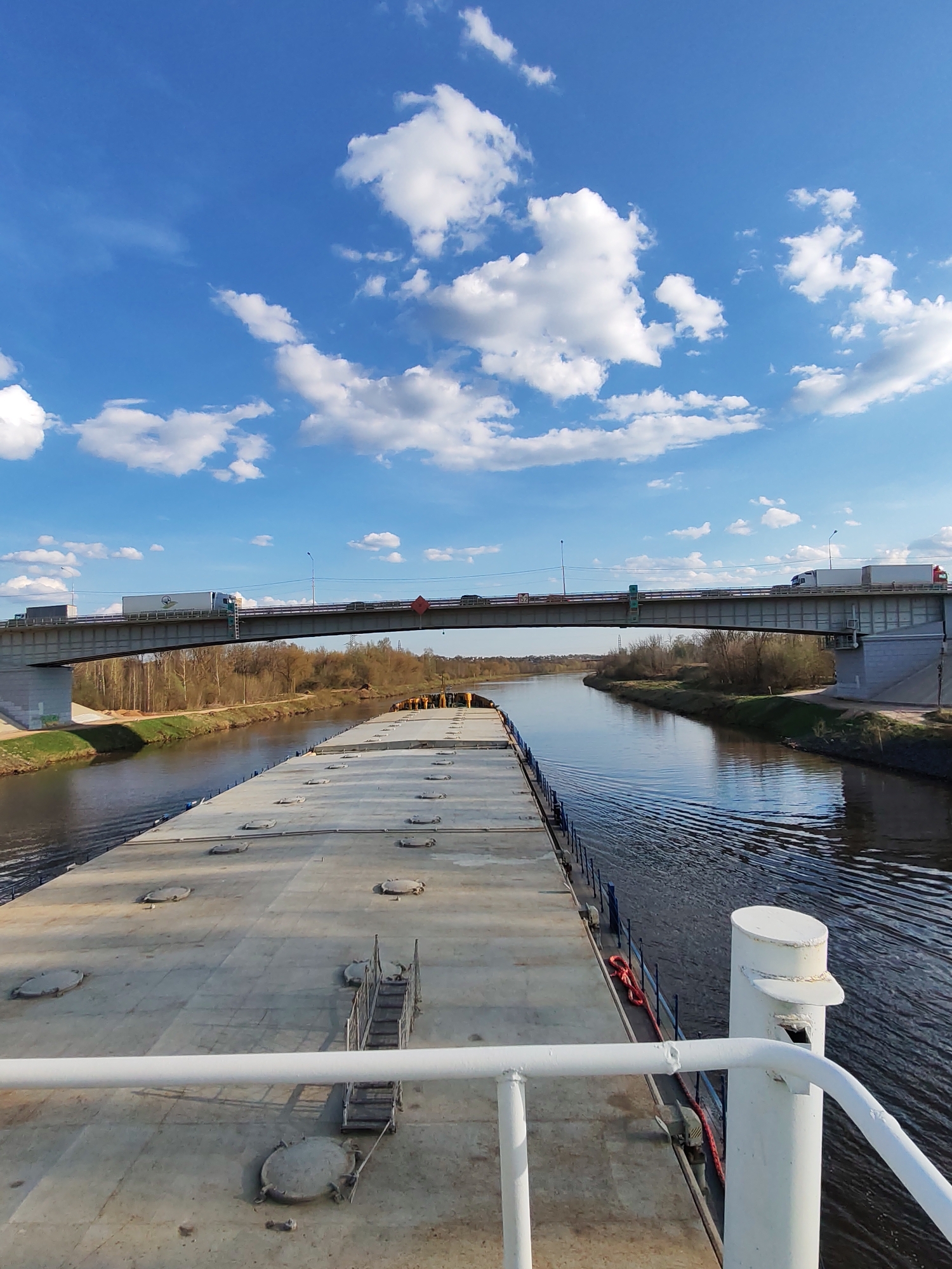 The image depicts a barge navigating a river in Podosinki, Russia, beneath a highway bridge. The barge deck is concrete and features multiple access hatches and a small staircase. The river reflects the blue sky with scattered clouds. The river banks are lined with trees and vegetation in early spring. The bridge carries vehicular traffic, including at least two large trucks. The viewpoint is from the front of the barge.