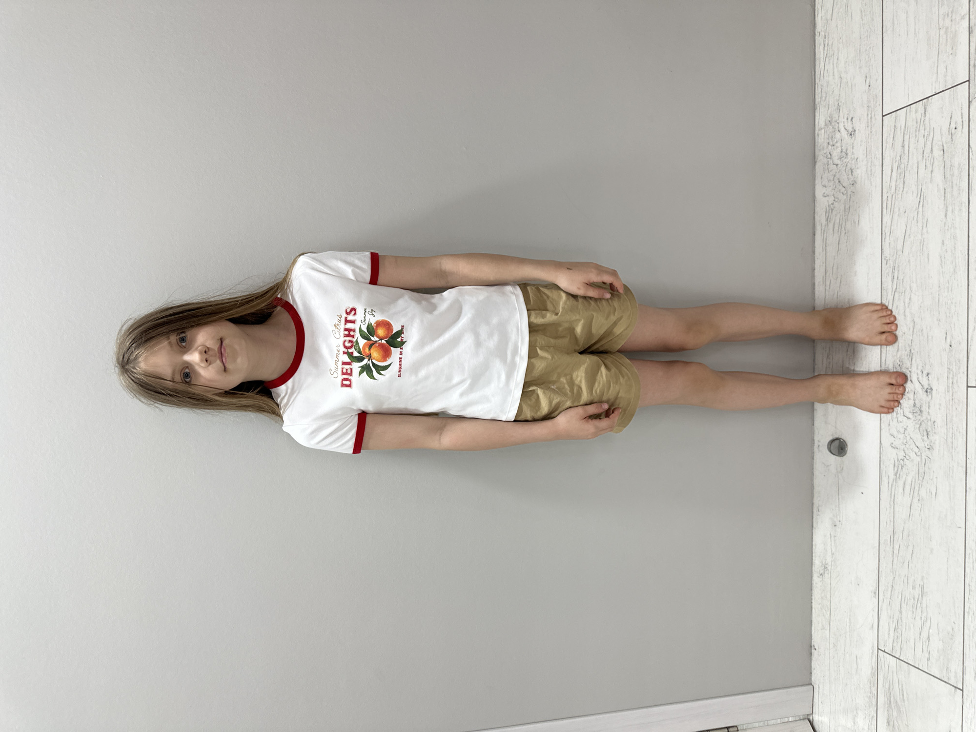 A young girl with shoulder-length blonde hair stands barefoot in front of a light grey wall, her expression neutral. She is wearing a white t-shirt with a red trim around the neckline and sleeves, featuring an image of oranges and the words "Summer Citrus DELIGHTS" in red lettering. Below the t-shirt, she wears light brown, loose-fitting shorts. The floor beneath her is tiled in a wood-grain pattern with greyish-white tones. A sliver of a white doorframe is visible on the left edge of the image. This scene is not depicted in the city of Sochi, Russia.