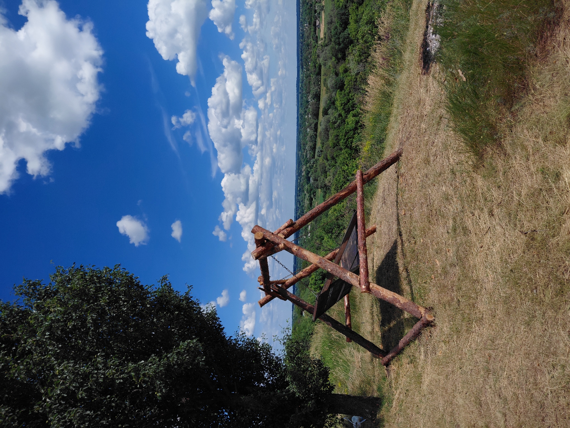 A wooden swing set with a hammock is situated on a grassy hillside overlooking a lush green landscape under a bright blue sky dotted with white clouds. To the left, a large tree with dark green leaves casts a shadow over part of the scene. The swing set is constructed from rough, natural wood logs and has a dark hammock stretched between two A-frame structures. The ground around the swing set is covered in dry, yellowed grass. In the background, rolling hills covered in dense green trees stretch towards the horizon, with a few scattered buildings visible in the distance. The overall scene suggests a peaceful, natural setting.