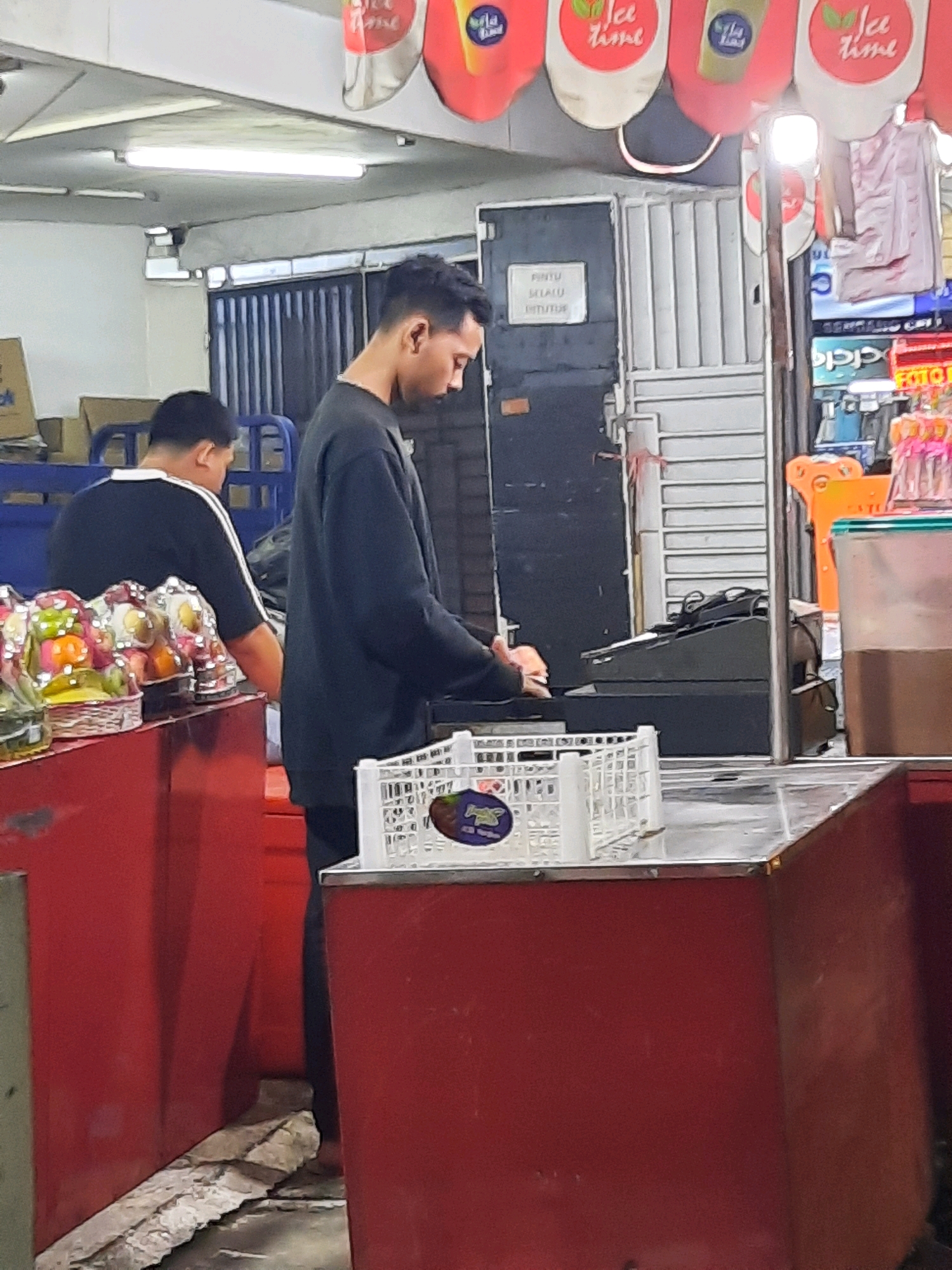 The image captures an indoor scene, likely a market or shop, in East Jakarta, Indonesia. In the foreground, two men are visible. The man on the right, with short, dark hair, is wearing a dark sweatshirt and is standing behind a red counter, seemingly engaged with items on the counter. To his left, another man, also with short dark hair, is wearing a black t-shirt with white stripes on the sleeves and is facing away from the camera, looking down at items on a red display counter.
In front of the man on the right, a white plastic crate is placed on the counter, containing some dark items and a sticker with a cartoon character. Behind him, a cash register is visible. The red display counter in the foreground holds several baskets of fruit, some of which are wrapped in clear plastic. The background reveals a metal door with vertical slats, a sign with text that appears to be in Indonesian, and various retail displays, including hanging decorations with "Ice Time" written on them, and shelves with products. The lighting appears to be artificial, with fluorescent lights visible on the ceiling. The overall environment suggests a commercial establishment.