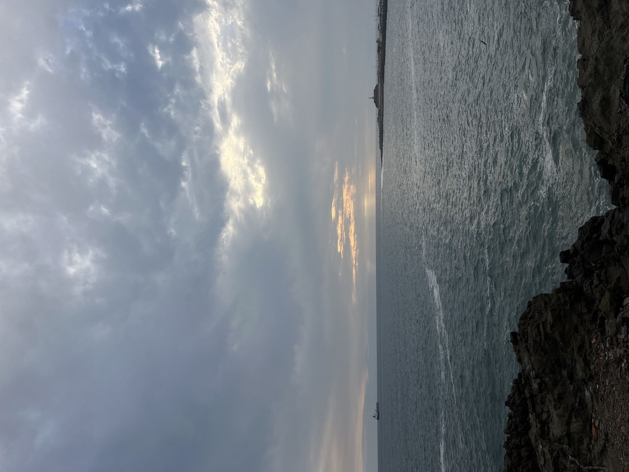 This photograph depicts a coastal scene likely in Arica, Chile, recognizable by its characteristic cliffs. The sky is overcast with a mix of gray and white clouds, with a hint of sunlight breaking through towards the horizon. The ocean stretches out from the right side of the frame, its surface rippled by waves. The foreground includes rocky formations.