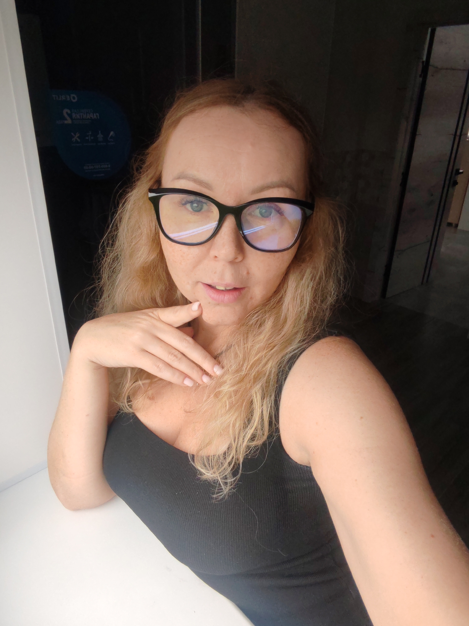 The image is a close-up, eye-level selfie of a woman with long, wavy, light blonde or light brown hair and fair skin with freckles. She is wearing black-rimmed glasses, through which her blue eyes are visible. Reflections of a light source are present on the lenses of her glasses. She is dressed in a black ribbed tank top or camisole.
The woman is looking directly at the camera with a neutral expression, possibly a slight smirk. Her right hand is gently placed near her chin and neck. Her left elbow rests on a plain white surface, which contrasts with the darker background.
The setting appears to be indoors. To the viewer's left, there is a dark window or glass panel that reflects a blue circular sign. To the viewer's right, the background is dark, suggesting a doorway or an opening to another dimly lit room.
On the reflected blue circular sign, several pieces of mirrored white text and symbols are discernible. The text includes "TIRJO" (likely "OJRIT" reversed) and "S RNTHARAT" (likely "TARHTANR S" reversed). Below these are four small icons, one of which appears to be an accessibility symbol (person in a wheelchair). Further down, a sequence of numbers and dashes is visible, resembling a phone number: "23-56-101-450-4". The overall lighting suggests it is daytime, with light illuminating the subject from the front.