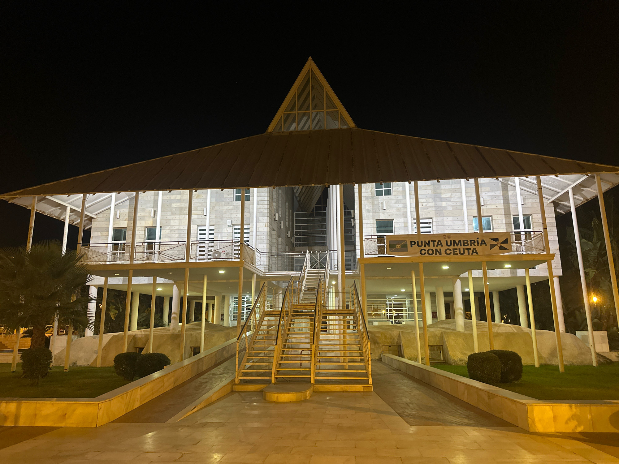 The image captures the exterior of a modern, multi-story building at night. The building features a prominent, angular, translucent roof structure extending over a wide, covered walkway. The facade is constructed of light-colored, textured blocks, with multiple windows on each level, some of which are illuminated. A central, open-air stairway, painted yellow and with metal railings, ascends to an upper level, flanked by ramps on either side. In front of the building, there is a paved walkway, illuminated by overhead lights, and a landscaped area with grass and shrubs, including a palm tree on the left. A banner with text and symbols, reading "PUNTA UMBRÍA CON CEUTA," is visible on the upper level of the building. The overall scene suggests a public or administrative building in Punta Umbría, Spain, possibly a civic center or government office, set against a dark night sky.