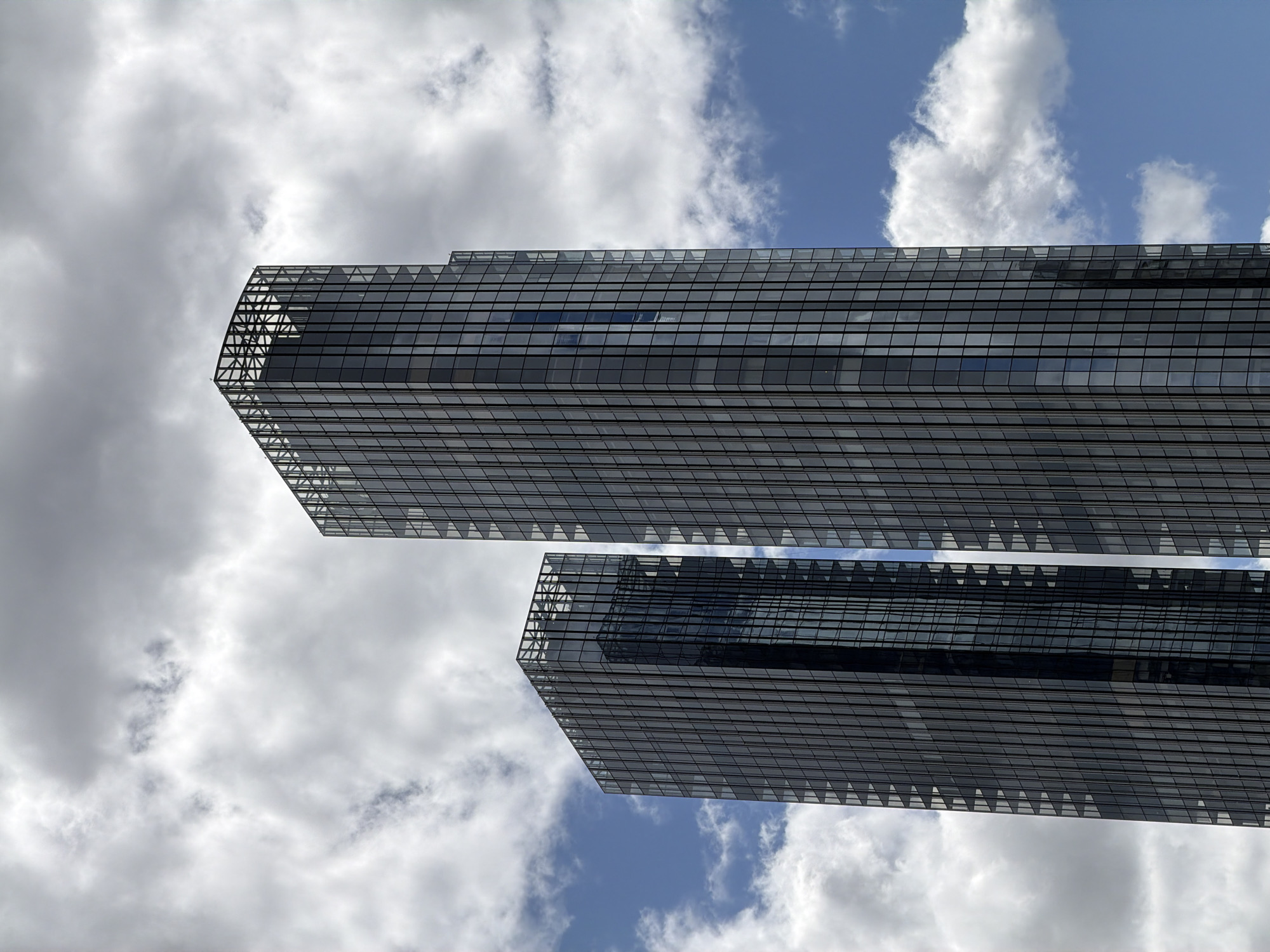 The image shows two modern skyscrapers against a partly cloudy sky. The buildings appear to have glass facades with a grid-like structure. The architecture features a distinctive top section with a triangular design element. The clouds are a mix of puffy white and darker gray formations. The buildings in this photo are located in the city of Moscow, Russia.
