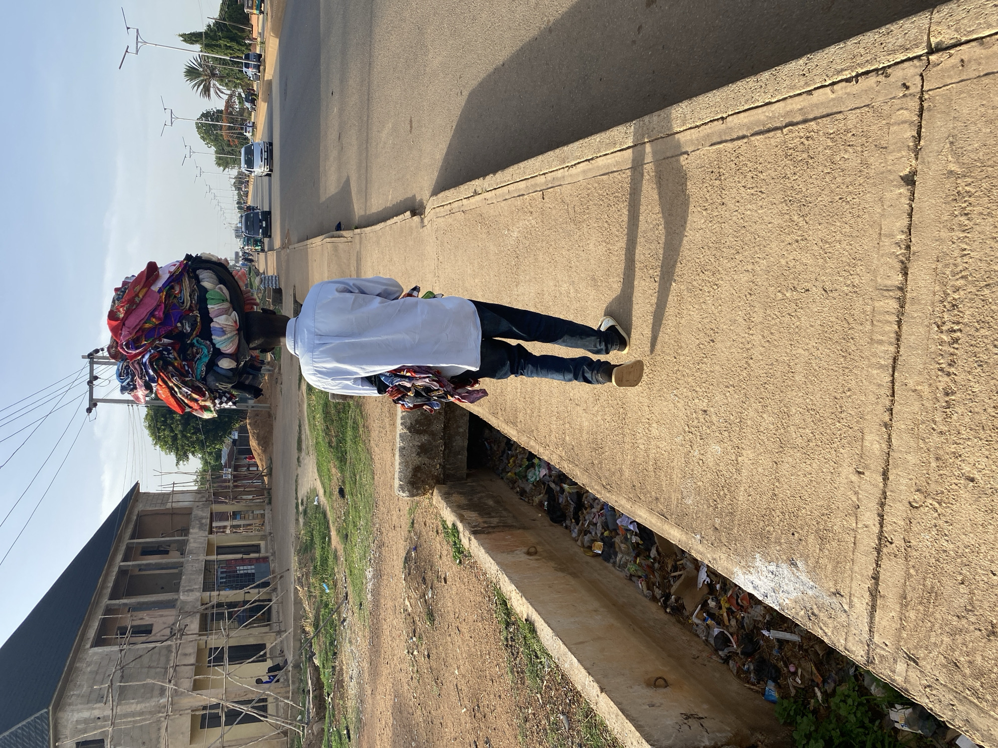 A street vendor is carrying a large bundle of colorful clothing on his head in Bukuru, Nigeria. He is walking along a concrete sidewalk next to a paved road with passing cars. Adjacent to the sidewalk is a ditch filled with trash. An unfinished building is visible across the ditch. The sky is clear and the sun is shining.