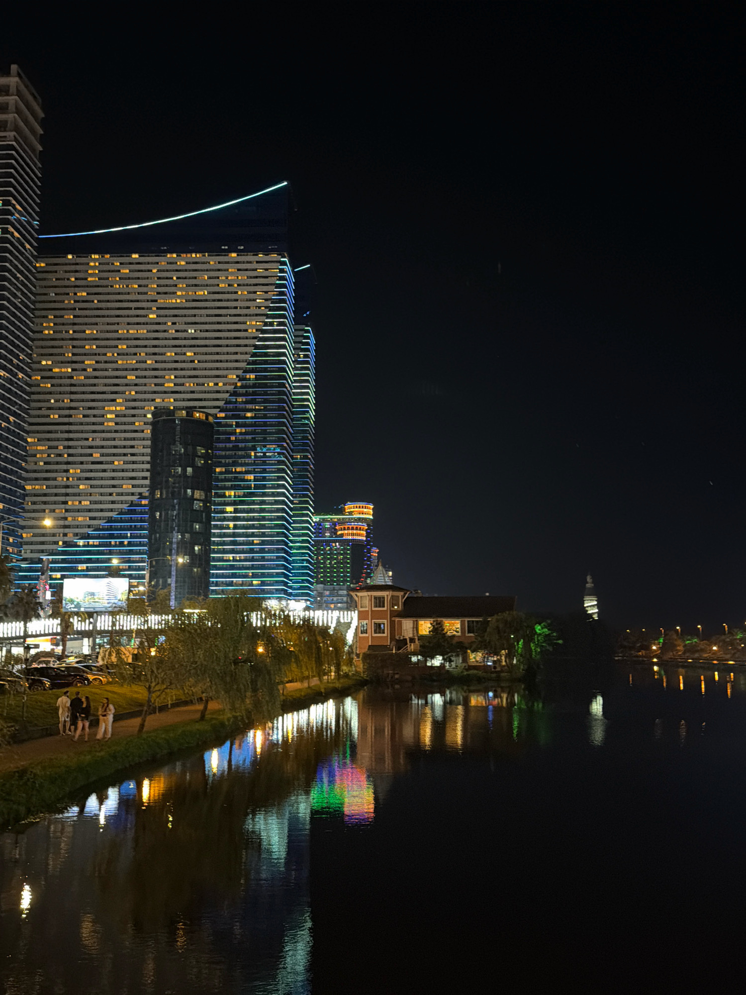 This is a nighttime shot of the Batumi Boulevard area in Batumi, Georgia, focusing on the waterfront and modern architecture. In the foreground, a dark body of water reflects the lights from the buildings and trees. Along the left bank of the water, a grassy slope leads to a paved walkway lined with trees, their branches casting shadows. Several people, appearing as small figures in the distance, are visible on the walkway, some standing and some walking. To the left of the walkway, a row of parked cars is dimly lit. Dominating the background is a cluster of contemporary high-rise buildings, illuminated with colorful LED lights. One building, particularly tall and slender, features horizontal bands of warm yellow lights, interspersed with vertical streaks of blue and green lights along its edges. Another building to its right showcases a similar pattern of colorful lights. Further in the distance, a more traditional building with a reddish-brown facade and illuminated windows sits closer to the water, surrounded by greenery. Beyond this, a spiral-shaped tower and a distant bridge with lights are visible. The overall scene depicts a vibrant urban waterfront at night.