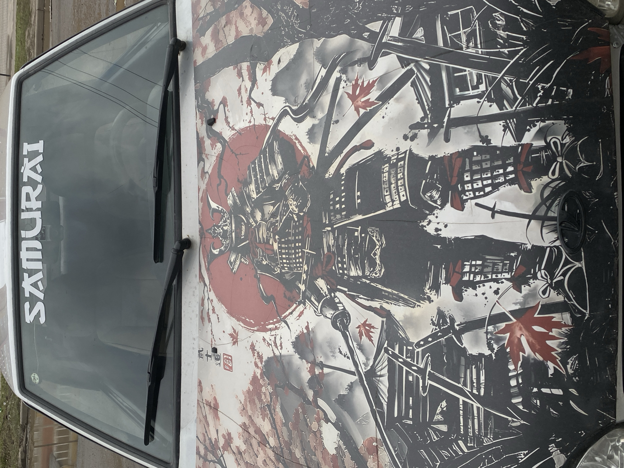 The image displays the front hood and part of the windshield of a white vehicle, featuring an intricate, custom artwork. The dominant subject is a detailed samurai-themed design covering the entire hood, rendered primarily in black, white, and red.
Central to the artwork is a large, weathered red circle, reminiscent of a Japanese rising sun, upon which is overlaid a depiction of a samurai helmet and armor, executed in a stylized, almost graphic novel aesthetic with sharp lines and shading. Surrounding this central figure are elements suggestive of traditional Japanese architecture, like castles or temples, and several red maple leaves and what appear to be cherry blossoms, adding splashes of color and a distinctive cultural motif. Swords, particularly katanas, are integrated into the complex background details. A small red square with black Japanese characters, possibly an artist's seal or signature, is visible on the hood near the windshield.
On the vehicle's windshield, a white vinyl decal vertically spells out the word "SAMURAI". Two black windshield wiper blades are also visible across the lower part of the windshield. The background visible through the windshield is blurred but indicates an outdoor setting, likely urban or suburban, with faint outlines of buildings or structures.
The vehicle appears to be parked during daytime, possibly under an overcast sky given the diffused lighting. The overall impression is a highly customized car reflecting an appreciation for Japanese warrior culture. The vehicle is located in Yoshkar-Ola, Russia.