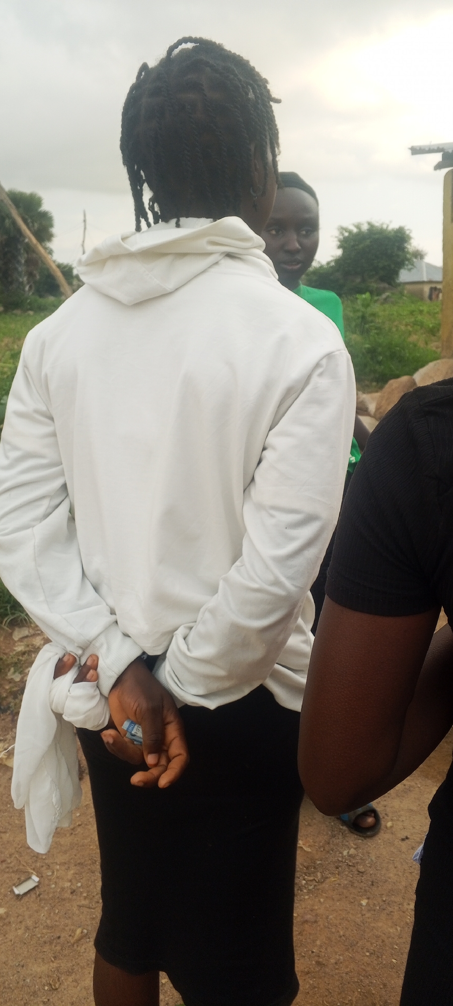 In Ohit, Nigeria, an individual stands with their hands behind their back, one hand tightly clutching a small wad of bills. They are wearing a white hooded sweatshirt and a black skirt. A white cloth is wrapped around their right wrist. Another person, wearing a green shirt, is visible in the background, looking towards the camera. The ground is composed of dirt and small stones. The sky is overcast.