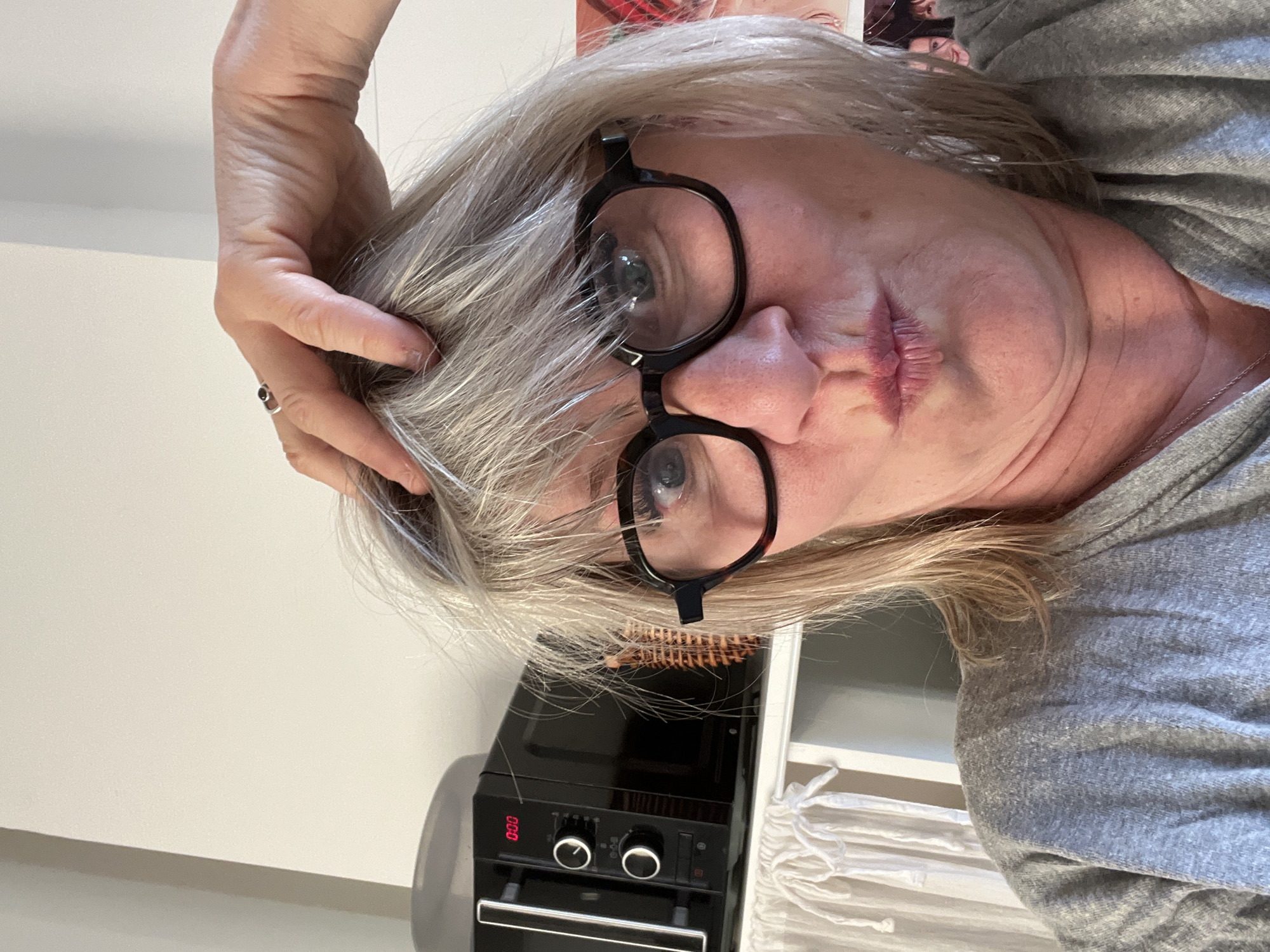 A woman with graying blonde hair and wearing large black-framed glasses is shown in a selfie-style photo. She is wearing a gray shirt and has her right hand placed on her head. A small oven is visible in the background, with the display showing "0:00". The location cannot be determined.