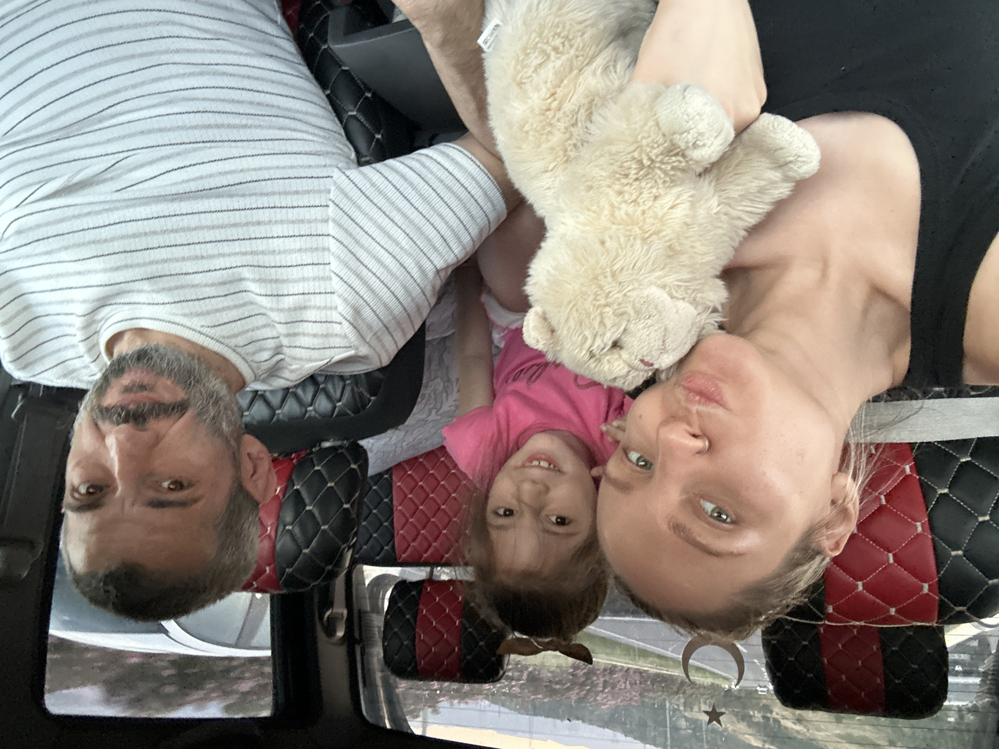 A family of three—a man, a woman, and a young girl—is in a vehicle. The man is on the left, wearing a striped shirt. The woman is on the right, wearing a black top and holding a stuffed animal bear. The girl, wearing a pink shirt, is between them. The vehicle's interior has quilted, red and black seats. Outside the window, the cityscape of Bornova, Turkey, is visible, with a decorative crescent moon and star visible on the window. The photo is taken from an overhead perspective.