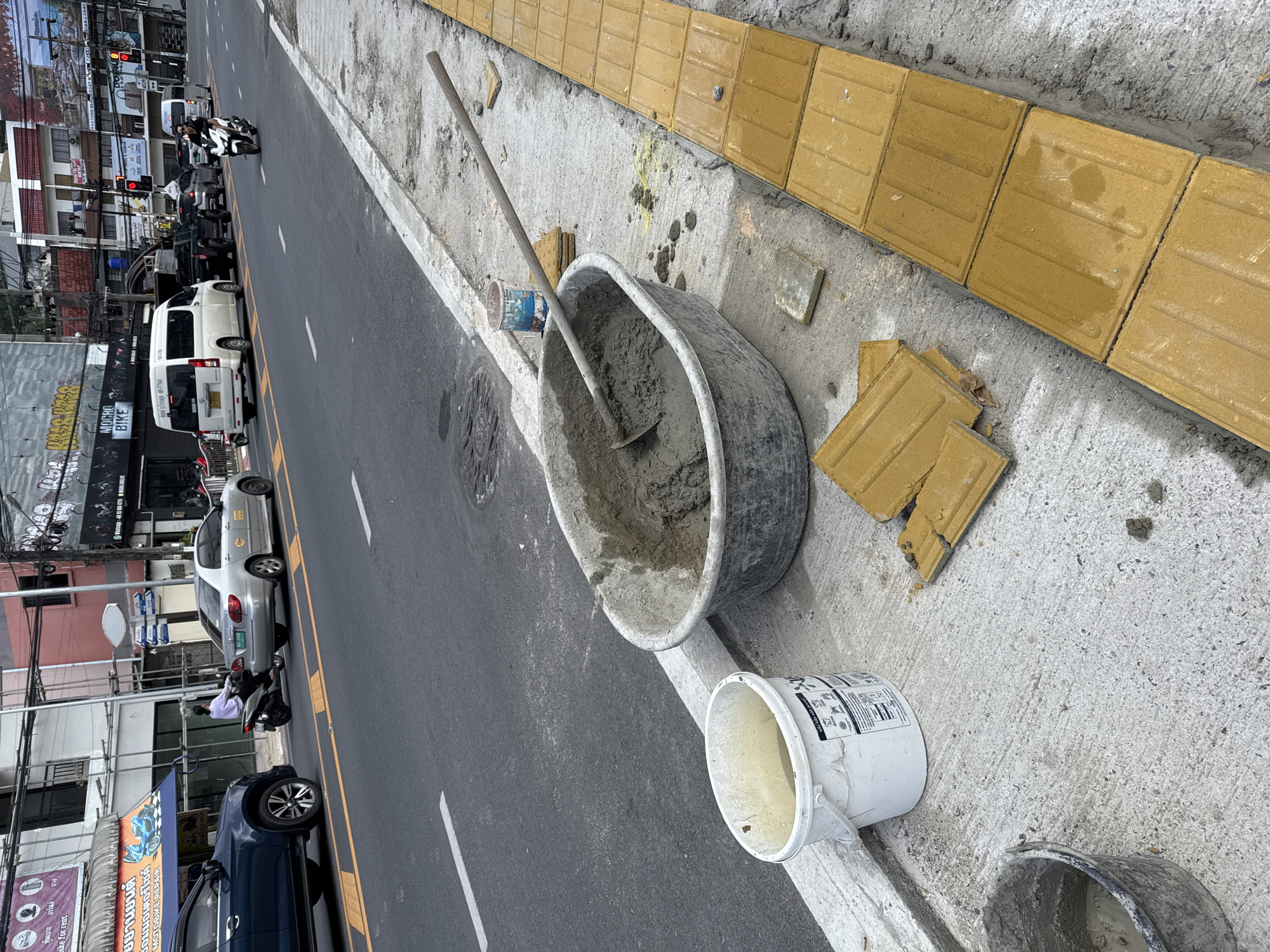 This is a street-level, eye-level shot of a construction scene on a sidewalk in Patong, Thailand. The foreground shows a large, gray mortar tub filled with wet cement, with a shovel partially submerged in it. To the left of the tub is a white plastic bucket with some water and residue inside. Further left, a black plastic bucket contains a small amount of cement. Stacked to the right of the mortar tub are several yellow tactile paving bricks, with more bricks laid in a row along the edge of the sidewalk. The sidewalk itself is made of concrete. In the background, there is a paved road with several vehicles, including a dark blue SUV, a silver sedan, a white van, and several motorcycles. People are visible riding motorcycles. Buildings line the street on the far side, with signs and wires suggesting a commercial area. The overall impression is a busy urban environment with ongoing sidewalk construction.