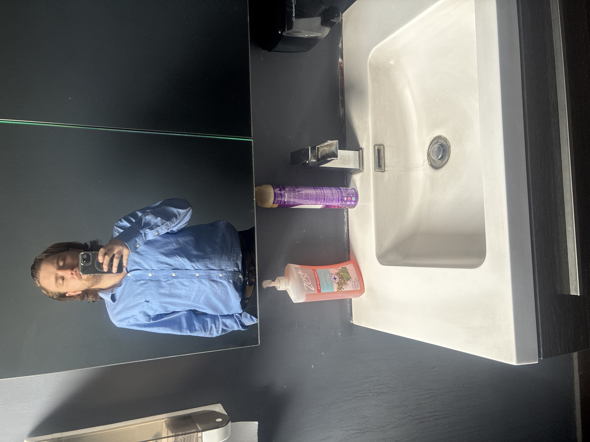 The image shows a bathroom sink with a white rectangular basin. There is a silver faucet and a bottle of Zest antibacterial hand soap to the side. A can of purple hairspray sits nearby. The background features a black countertop and a mirror. The reflection in the mirror shows a person holding a phone, wearing a blue button-down shirt and dark pants. It is impossible to verify the location in Cuautitlán Izcalli, Mexico without additional information.