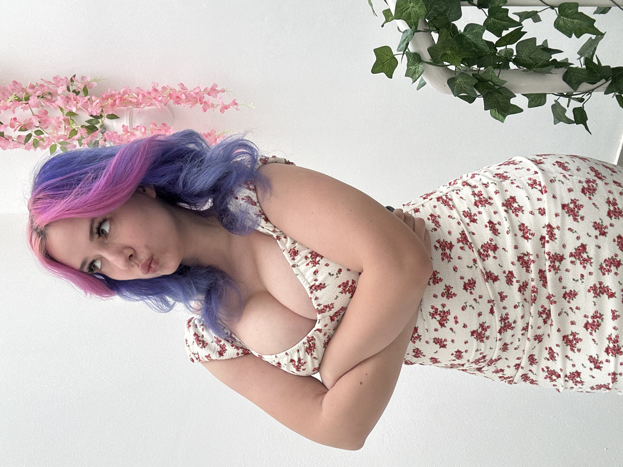 A young woman with distinctive purple and pink ombre hair is depicted in a tilted, close-up portrait. She is wearing a white dress with an all-over pattern of small red floral prints. Her arms are crossed over her chest, and she is looking towards the viewer with a pouting expression. Her eyes are visible, framed by dark eyebrows.
The background is a plain white wall. On the upper left side, a string of artificial pink cherry blossoms is draped, providing a soft, decorative element. On the upper right, a section of artificial green ivy leaves is visible, appearing to hang from a white support structure. The setting suggests an indoor environment, likely a studio or a brightly lit room. The woman's pose and playful facial expression are central to the image. This media file was created in the City of Zagreb, Croatia. There is no visible text in the image.